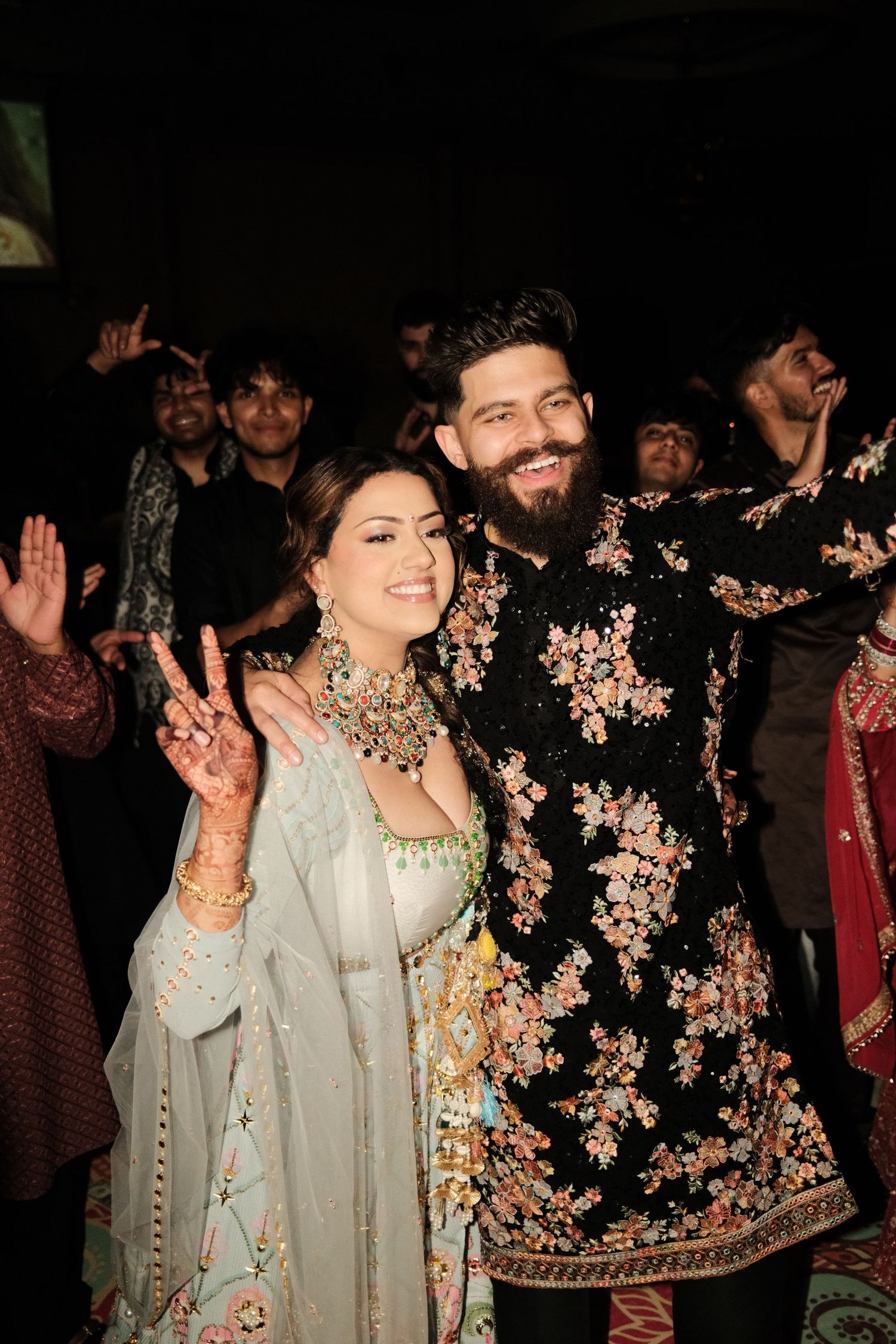 Smiling bride in a pastel lehenga with statement jewelry poses with groom in a black floral embroidered sherwani at a festive celebration.