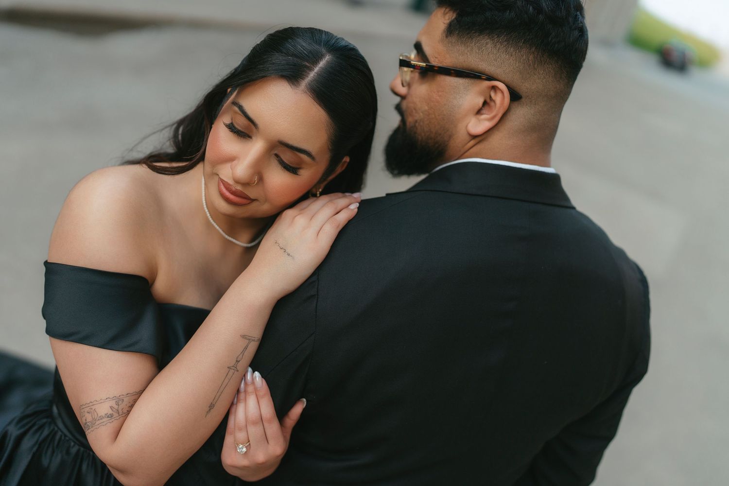Bride in a black off-the-shoulder gown rests her head on her groom's shoulder during a romantic engagement photo session.