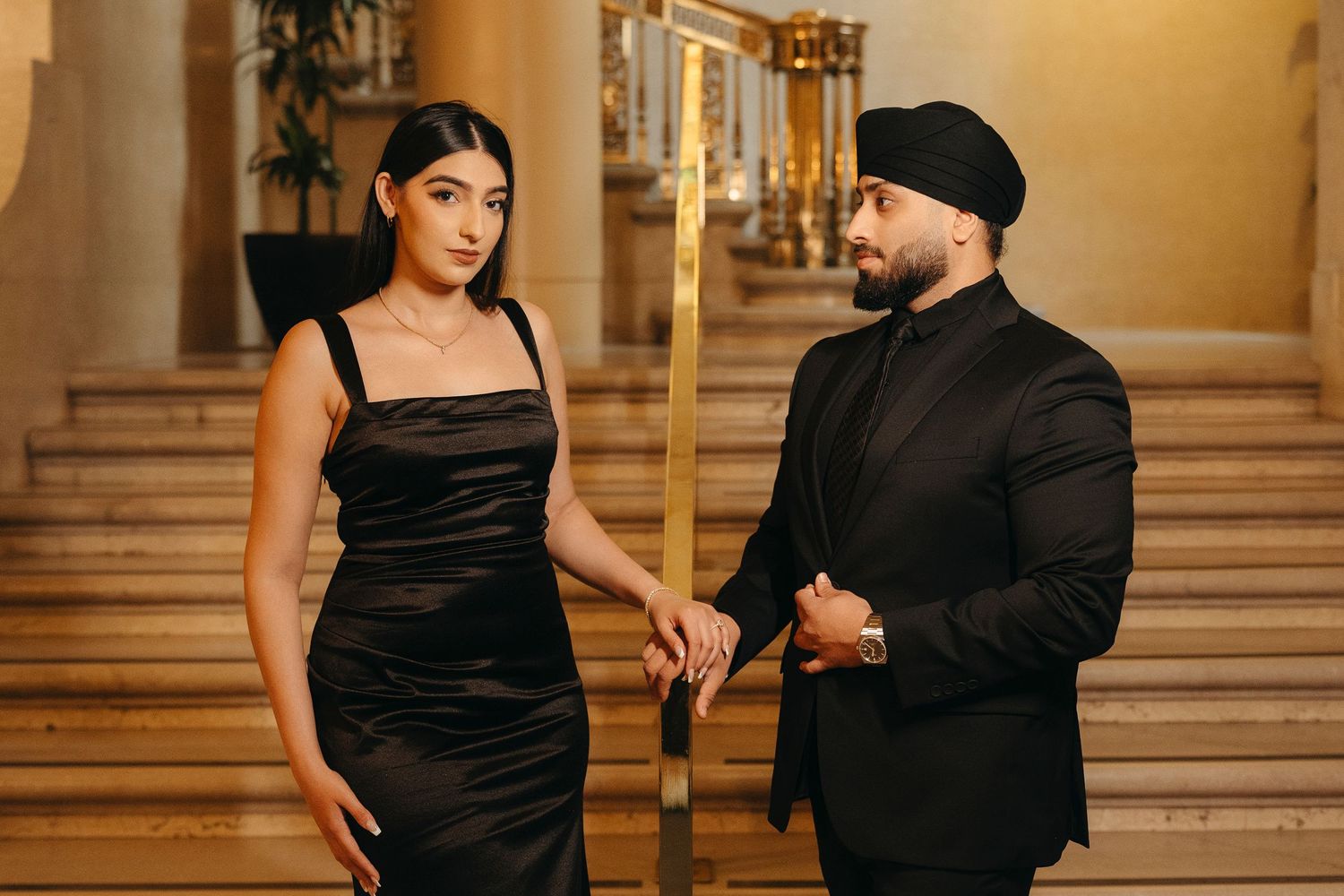 Couple in black formal attire hold hands on a grand staircase with golden railings during an engagement photoshoot.