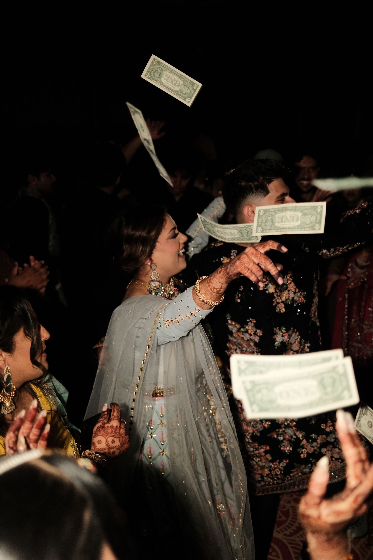 Dollar bills shower over a smiling bride and groom in traditional Indian attire as guests celebrate at a sangeet.