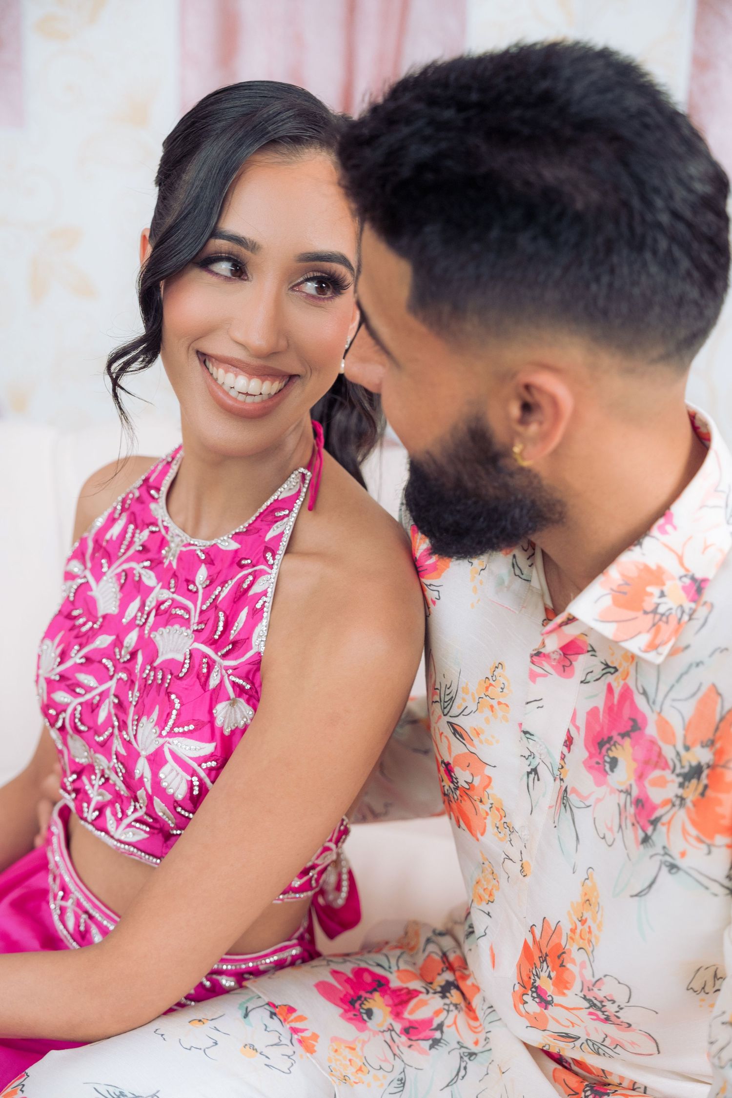 Smiling woman in a pink embroidered halter top gazes warmly as her bearded partner in a floral shirt leans close in a portrait.