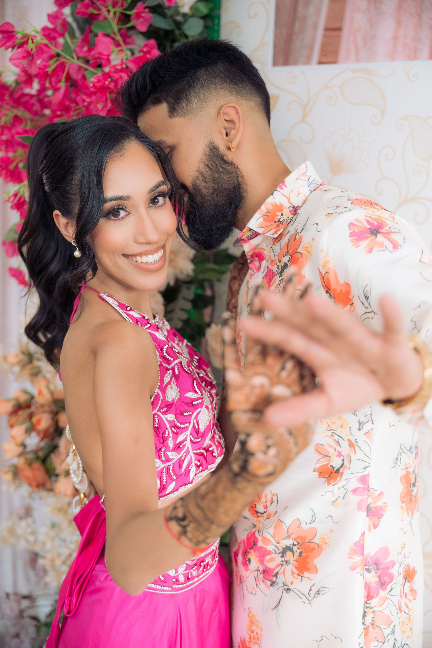 Couple in coordinated pink and floral outfits embrace while the bride shows off her intricate henna design beside pink blossoms.