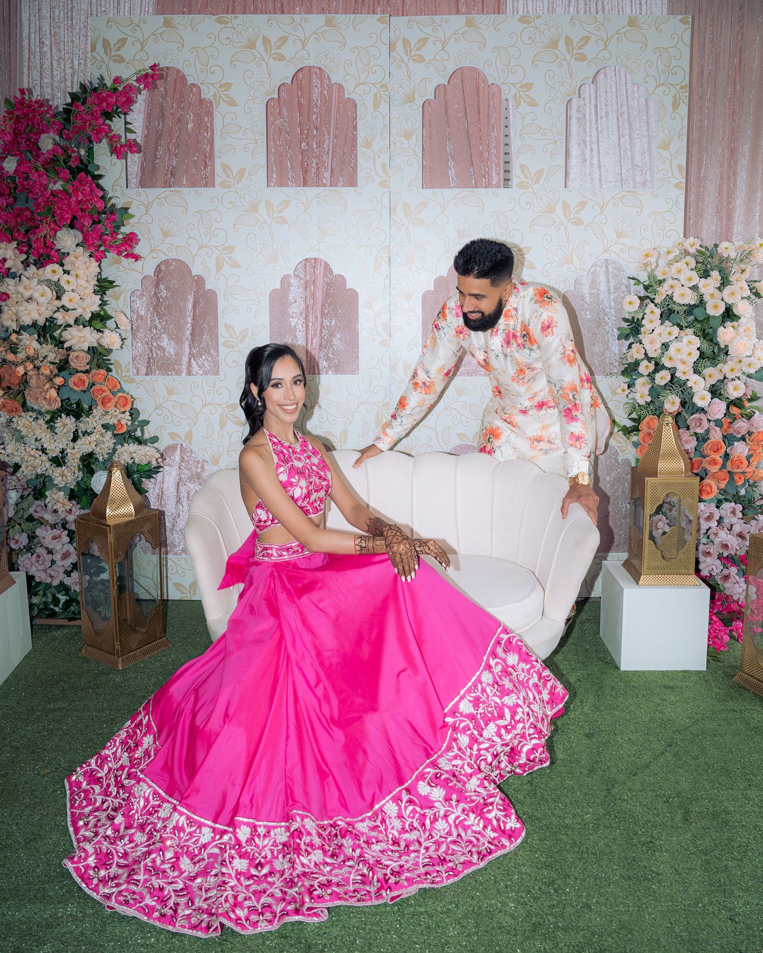 Bride in a vibrant pink embroidered lehenga sits on a white sofa as the groom in a floral shirt stands behind among flowers.