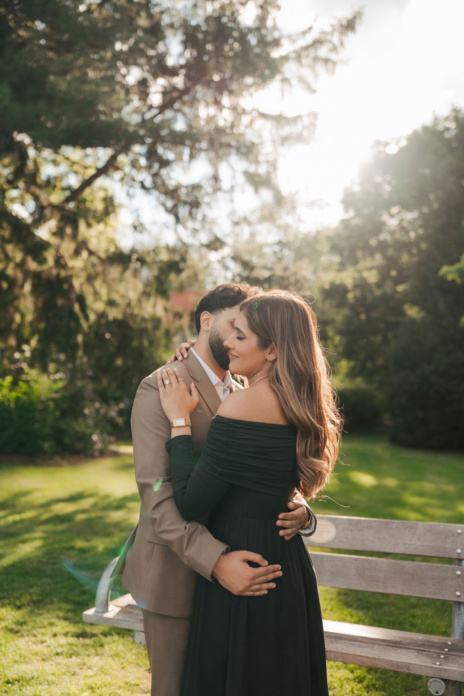 Couple embracing on a park bench in warm golden sunlight surrounded by green trees during an outdoor engagement photoshoot.