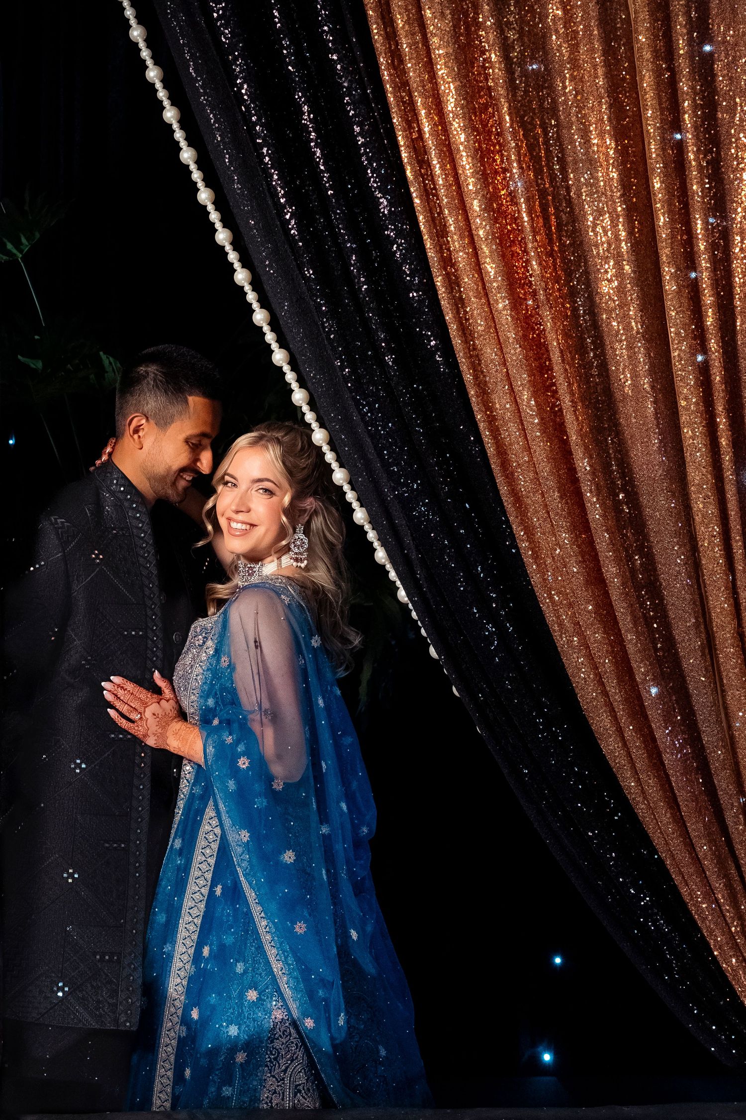 Smiling bride in a blue embroidered lehenga with jewelry leans into her groom in front of sparkling copper and black sequin curtains.