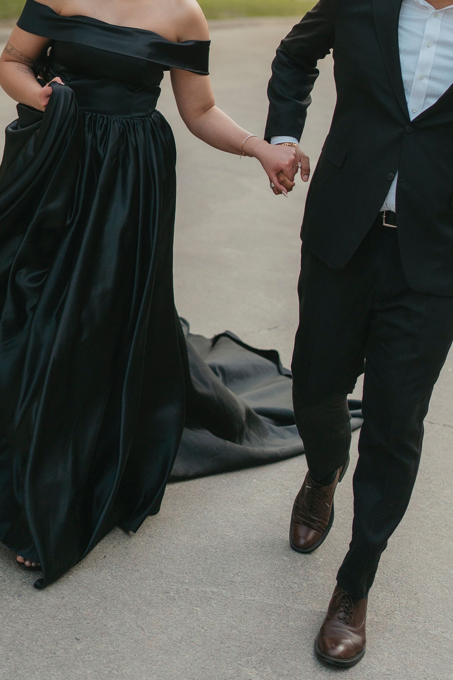 Close-up of a couple holding hands while walking, showing her flowing black satin gown with train and his black suit and brown shoes.