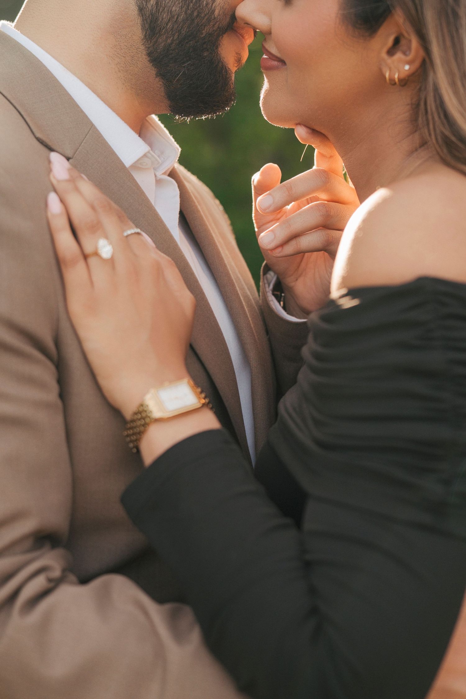 Close-up of a couple in a tender embrace, showing a sparkling engagement ring and gold watch, in warm natural light.