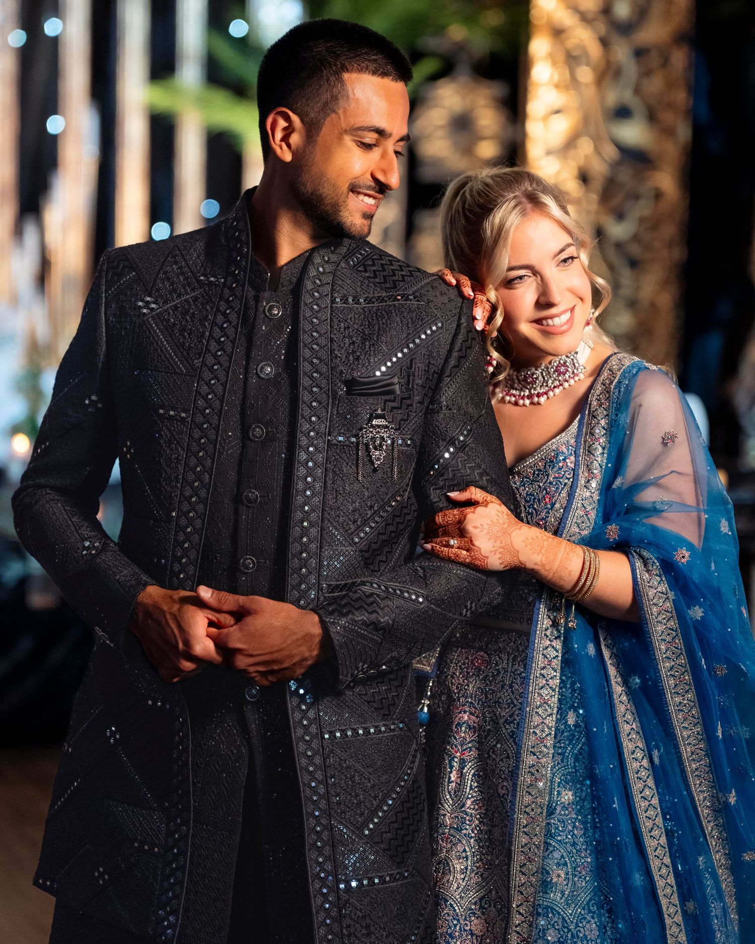 Smiling couple in coordinated Indian wedding attire, the man in an embroidered black sherwani and the woman in a blue lehenga with dupatta.