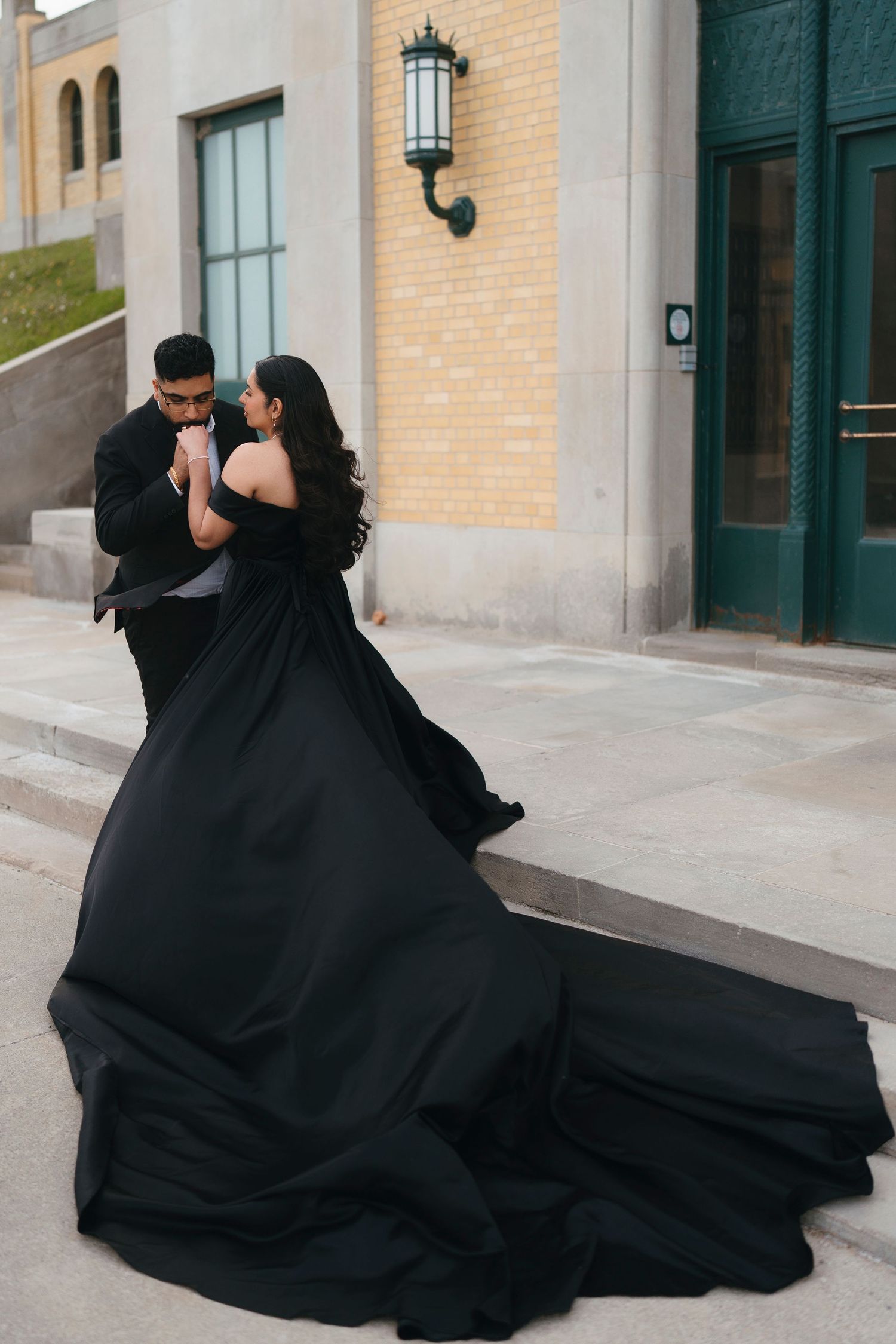 Couple embracing on stone steps, the bride in a flowing black ball gown with a long train and the groom in a black suit.