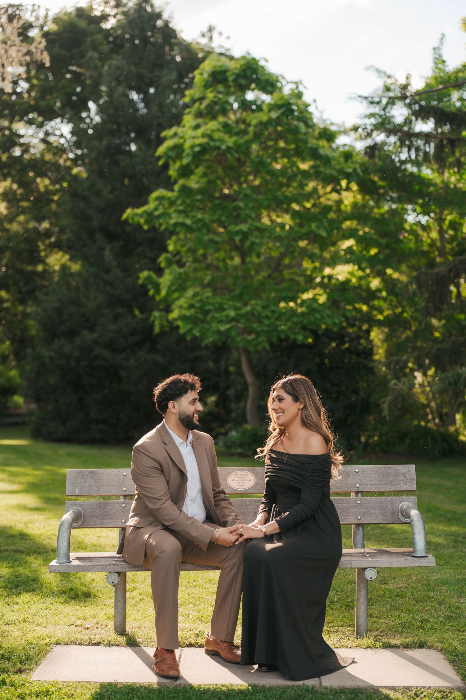 Engaged couple holds hands on a park bench beneath tall green trees on a sunny day.