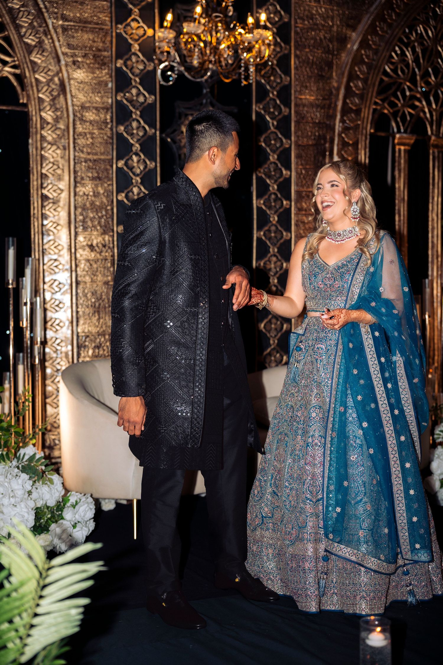 Groom takes his bride's hand as she laughs in a blue embroidered lehenga, set against ornate golden arches and a hanging chandelier.
