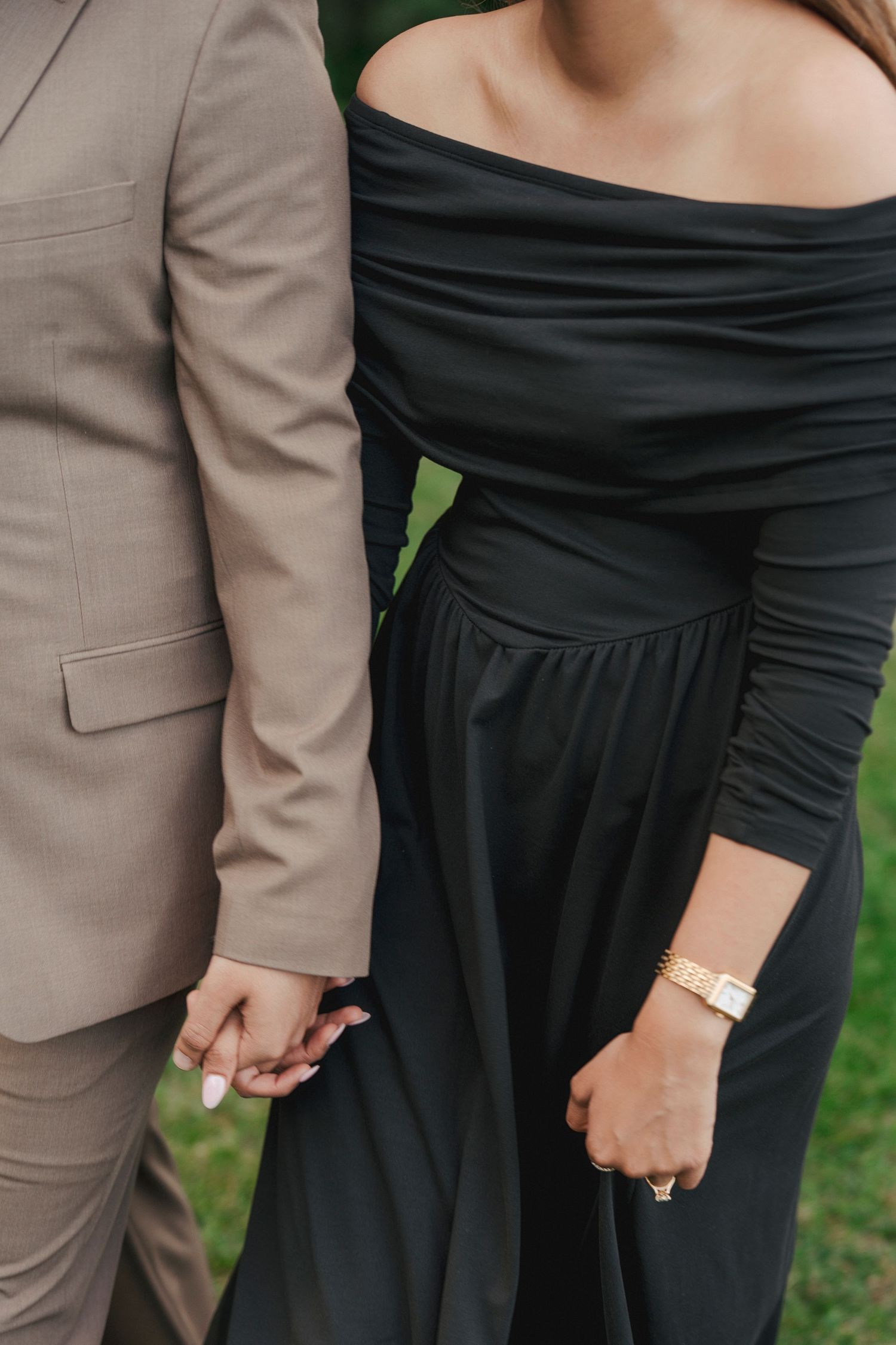 Couple holding hands on a lawn, she in a black off-shoulder dress with a gold watch, he in a tan blazer, in an engagement photo.