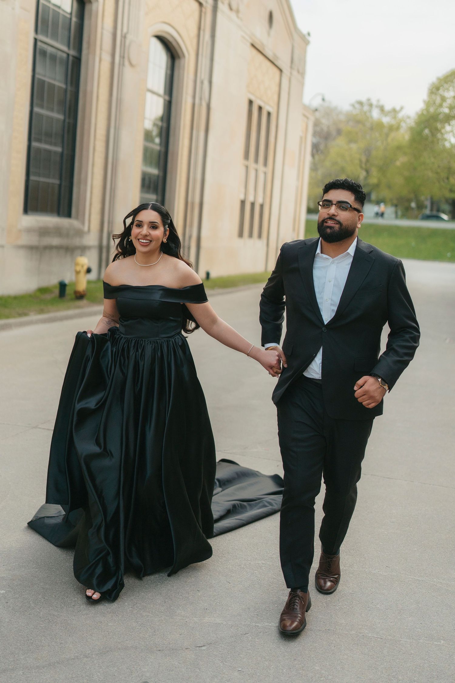 Smiling bride in a black satin off-the-shoulder gown holds hands with her groom in a dark suit as they walk outdoors.