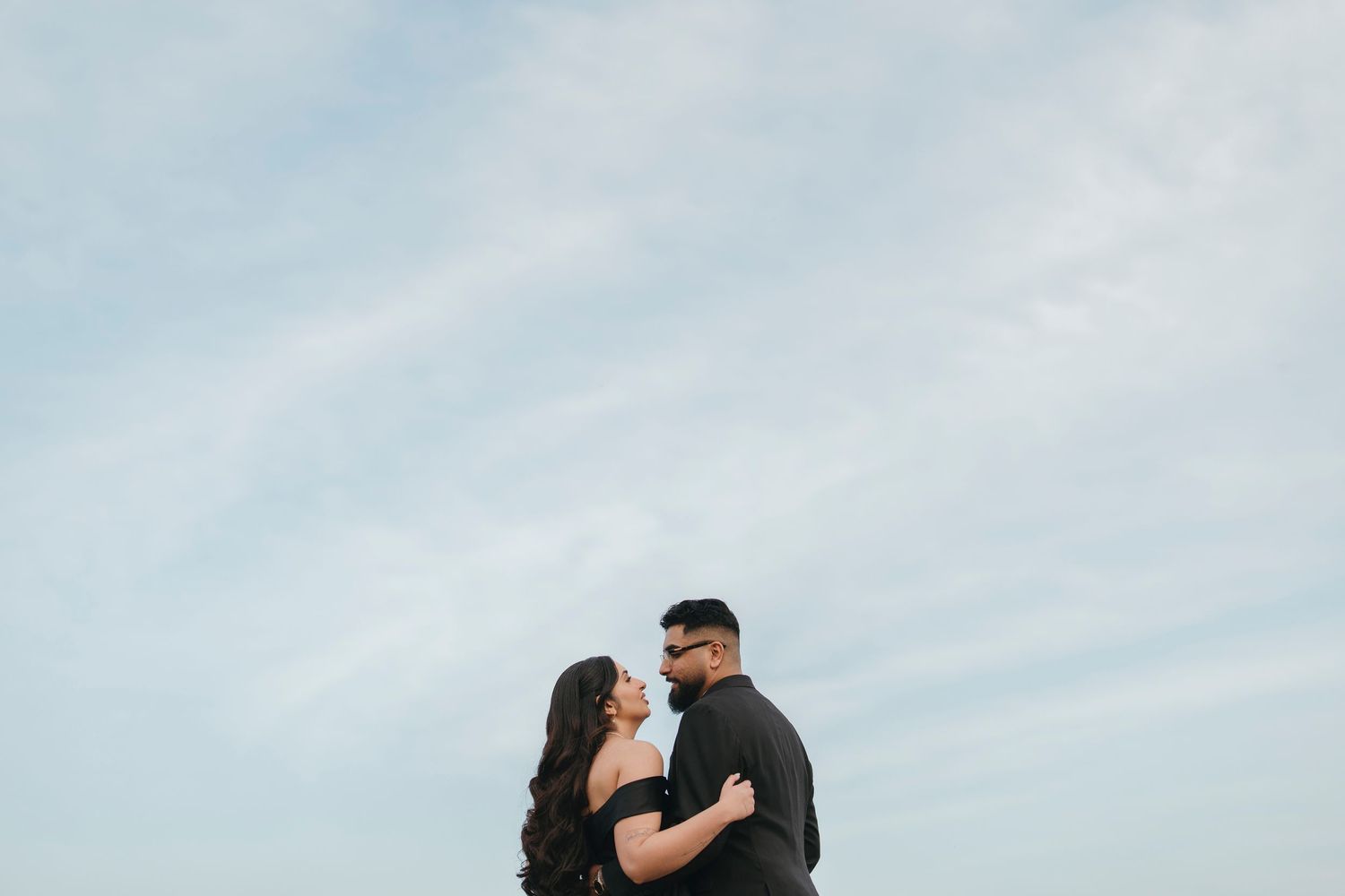 Engaged couple in black outfits share an intimate moment against a soft blue sky with wispy clouds.