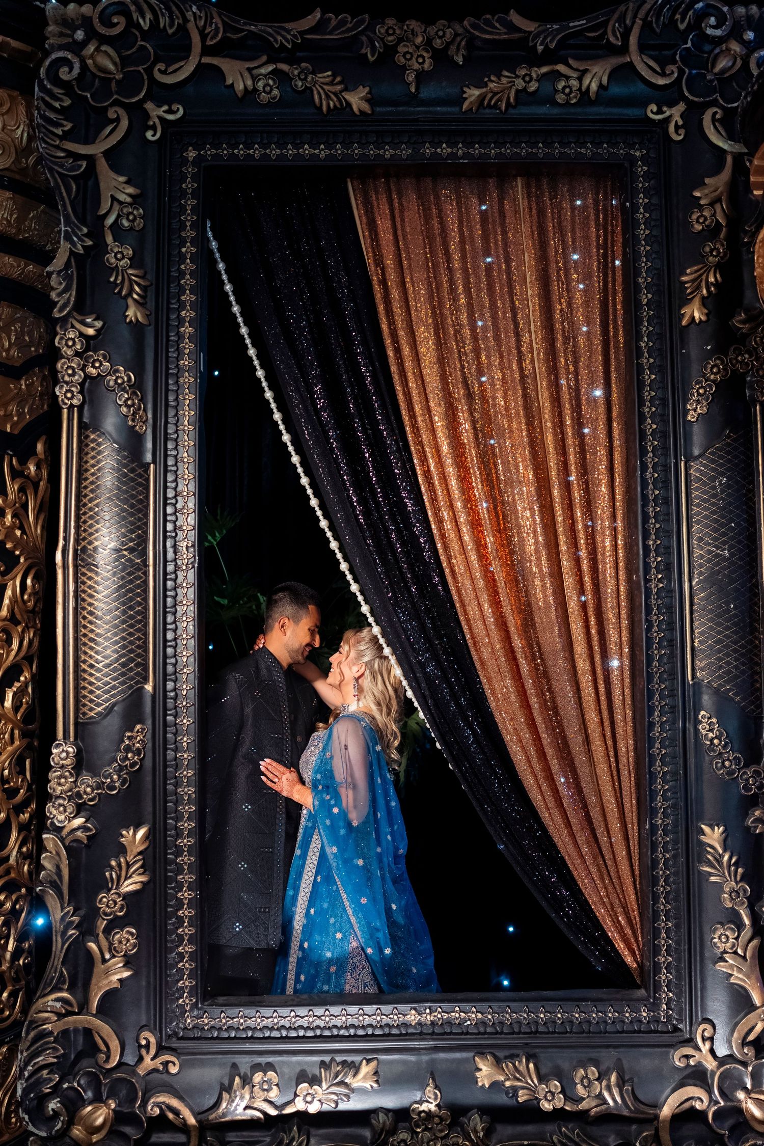 Couple embrace in an ornate black frame with shimmering copper and black sequin curtains, wearing black sherwani and blue lehenga.