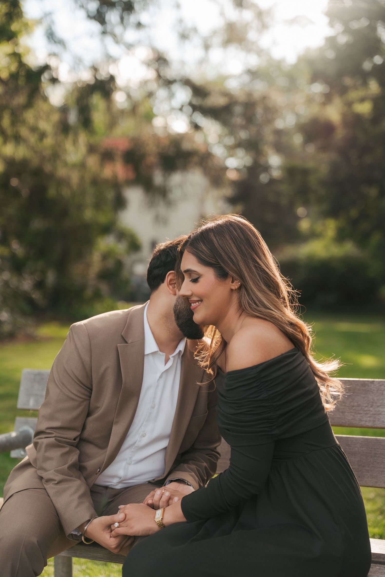 Smiling woman in a black off-shoulder dress shares a tender moment with her fiancé on a sunlit park bench.