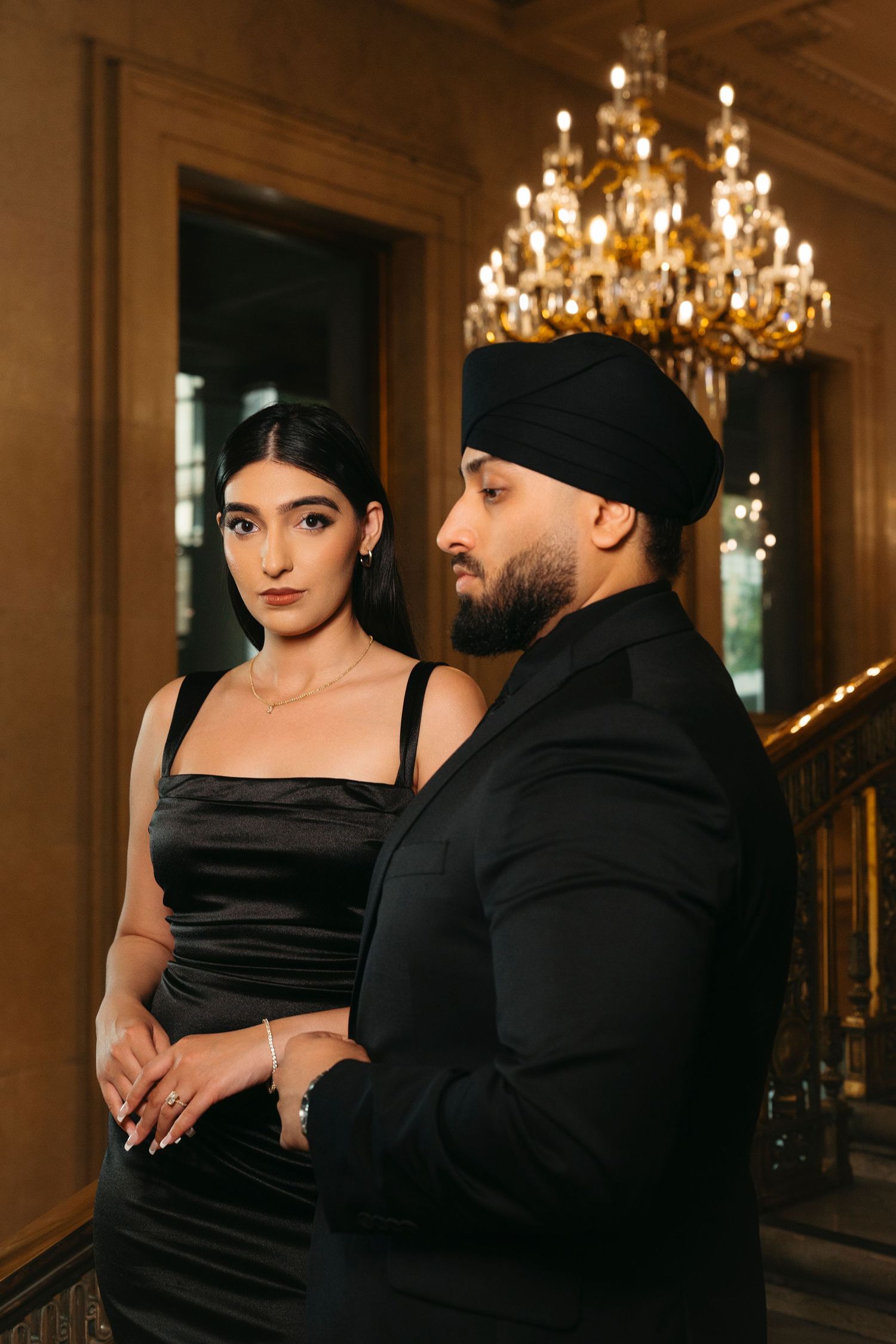 Elegant couple in black stand together beneath a glowing crystal chandelier in an ornate historic hall.