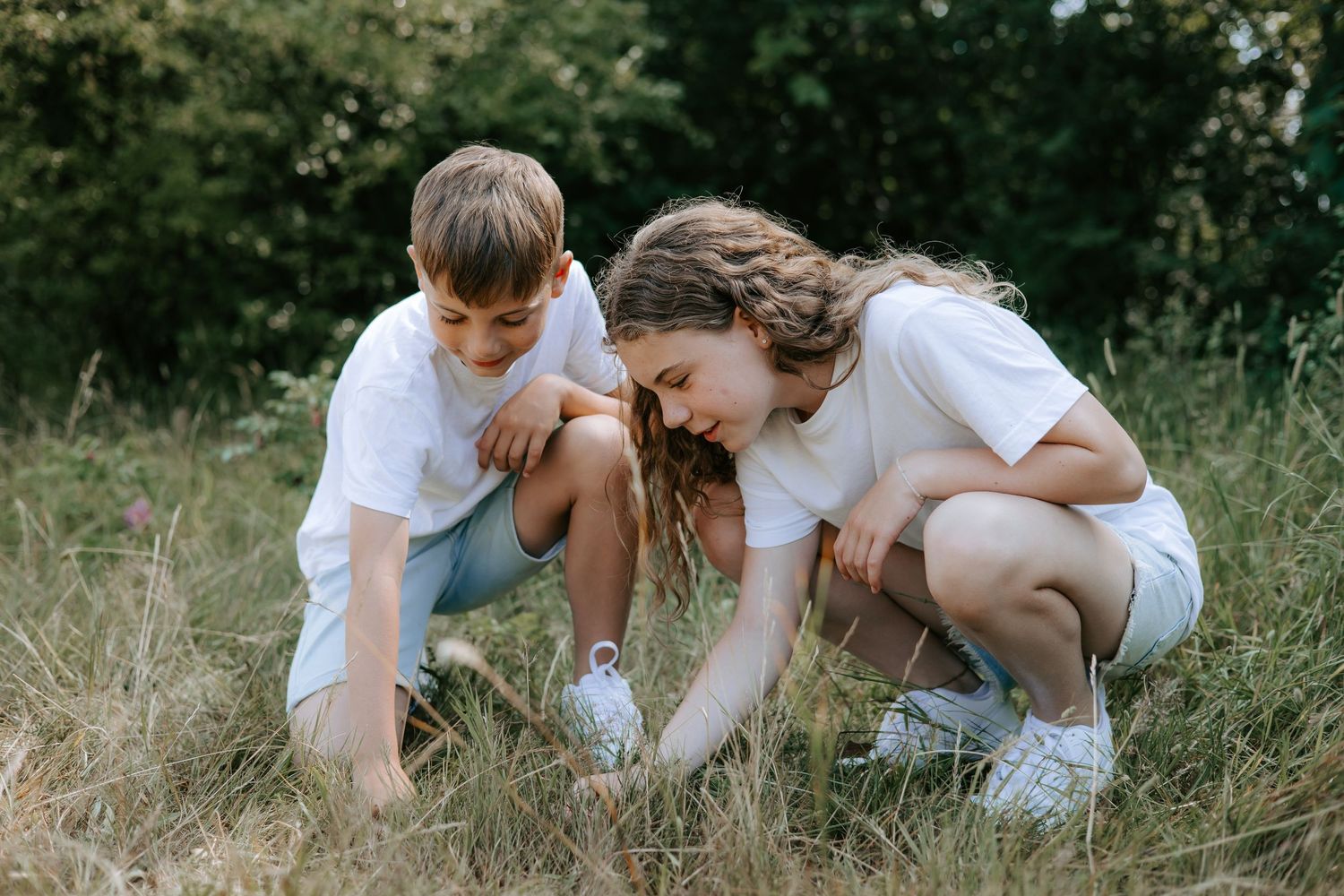 Two children crouching in grass examining something in nature on sunny day.