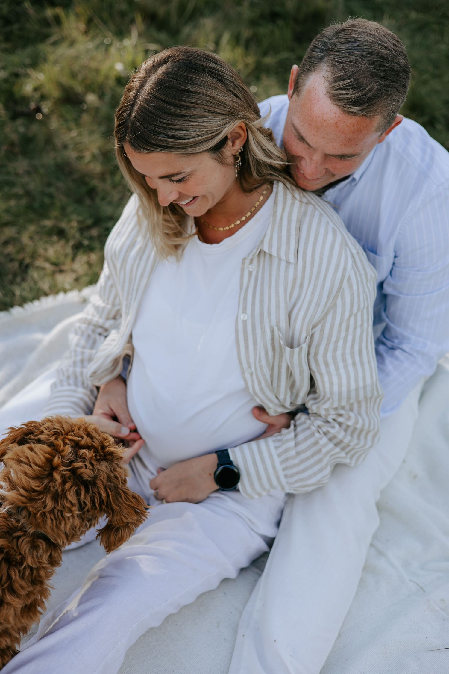 Pregnant couple sitting on white blanket outdoors, smiling as they cradle baby bump with a fluffy brown dog nearby.
