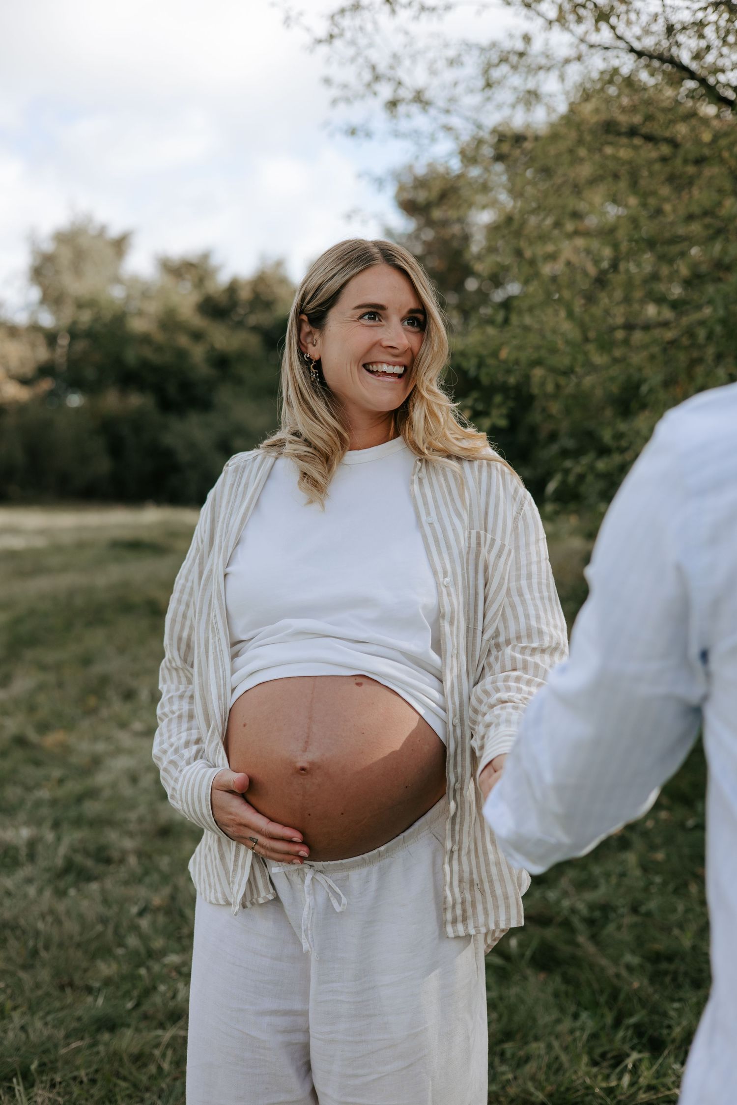 Smiling pregnant woman in white outfit and striped cardigan cradling her bare baby bump outdoors in a green field.