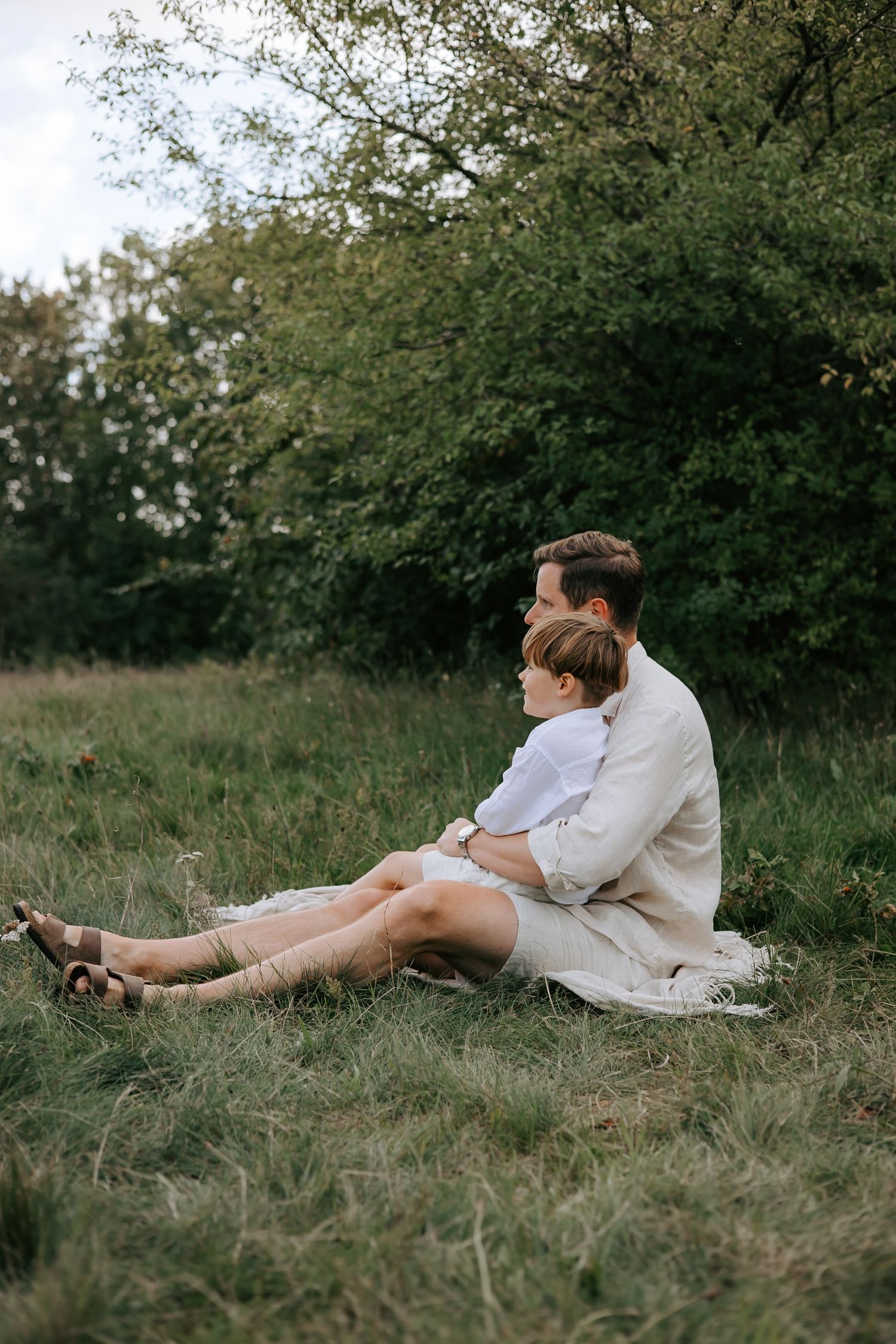 Father and young son sitting together on grass blanket in green field, sharing a quiet moment.