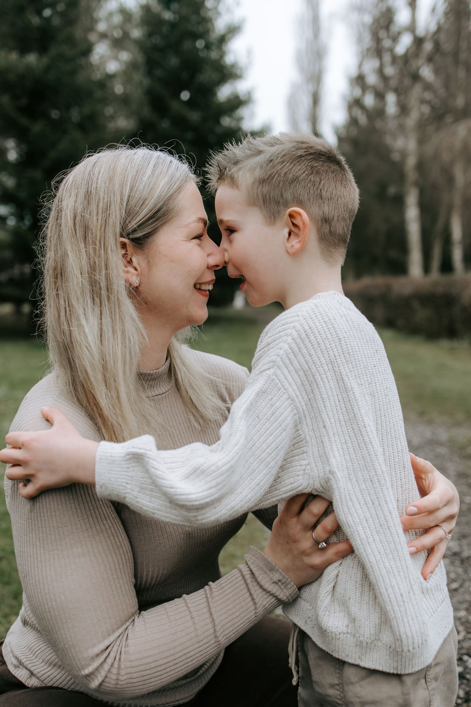 A smiling mother and young son touching foreheads and embracing affectionately in an outdoor park setting.