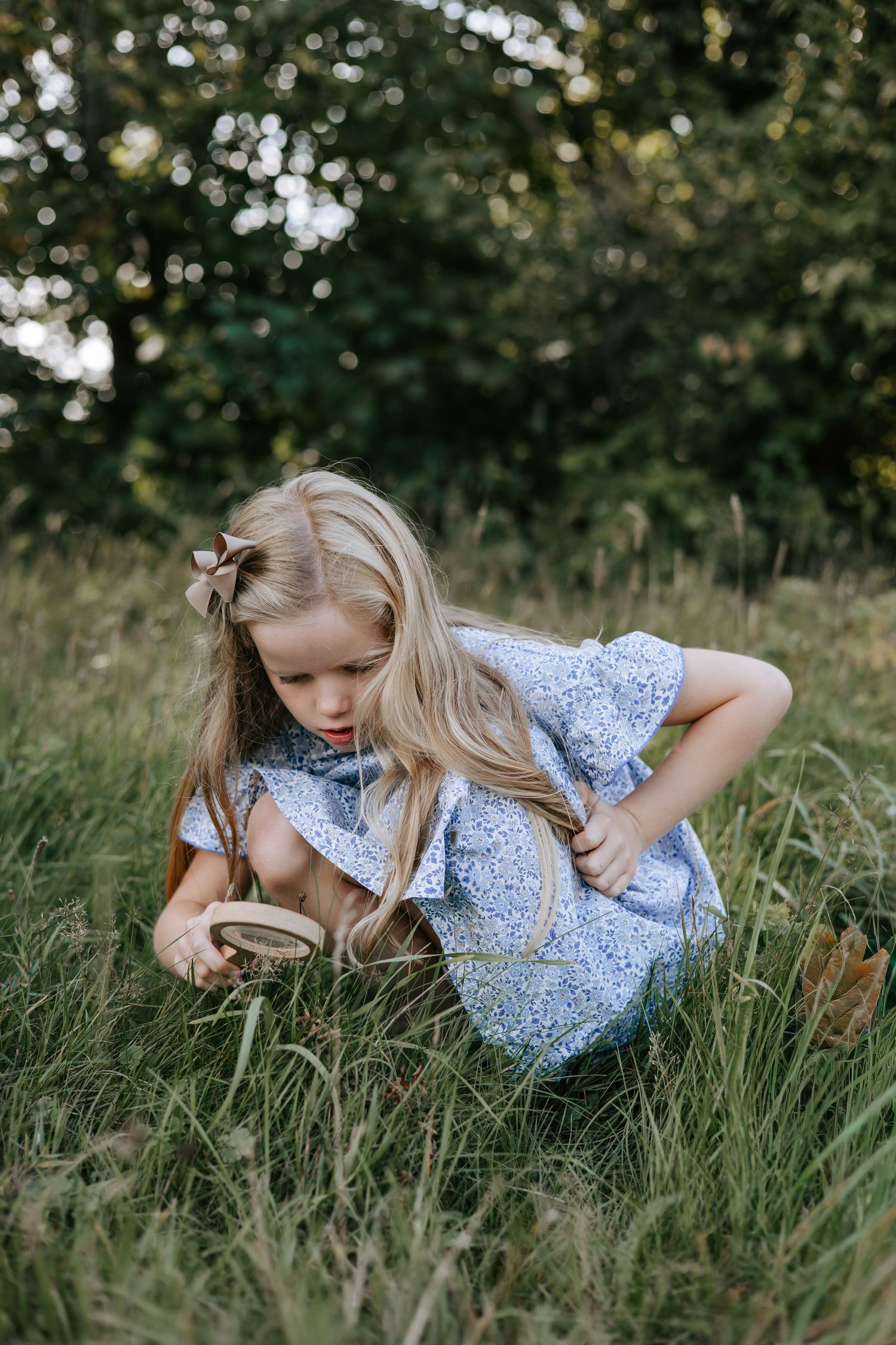 Curious blonde girl in blue floral dress examining grass with a magnifying glass outdoors.