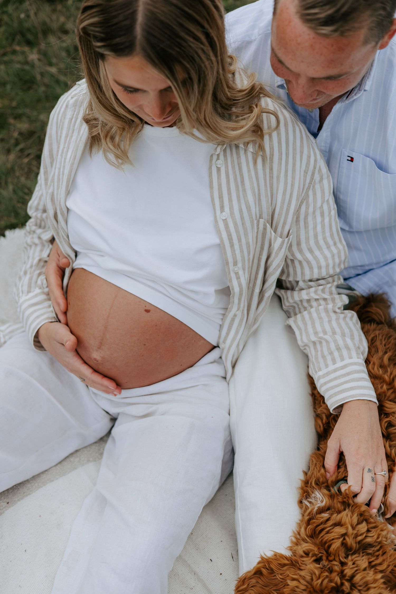 Pregnant woman in white outfit sits outdoors with partner, both gazing at her baby bump, with a fluffy dog nearby.