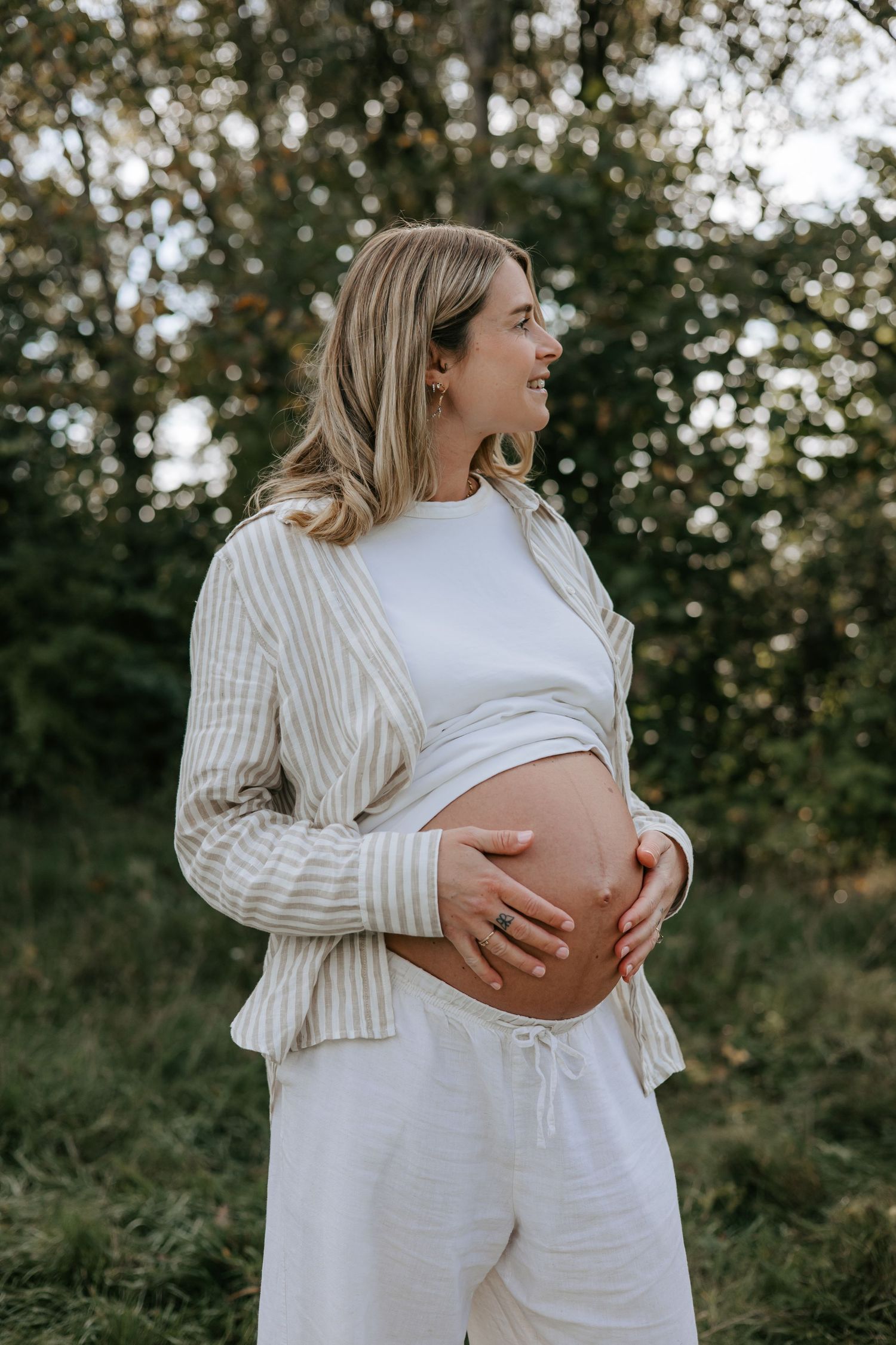 Pregnant woman in white outfit and striped cardigan cradling her baby bump outdoors in a lush green setting.