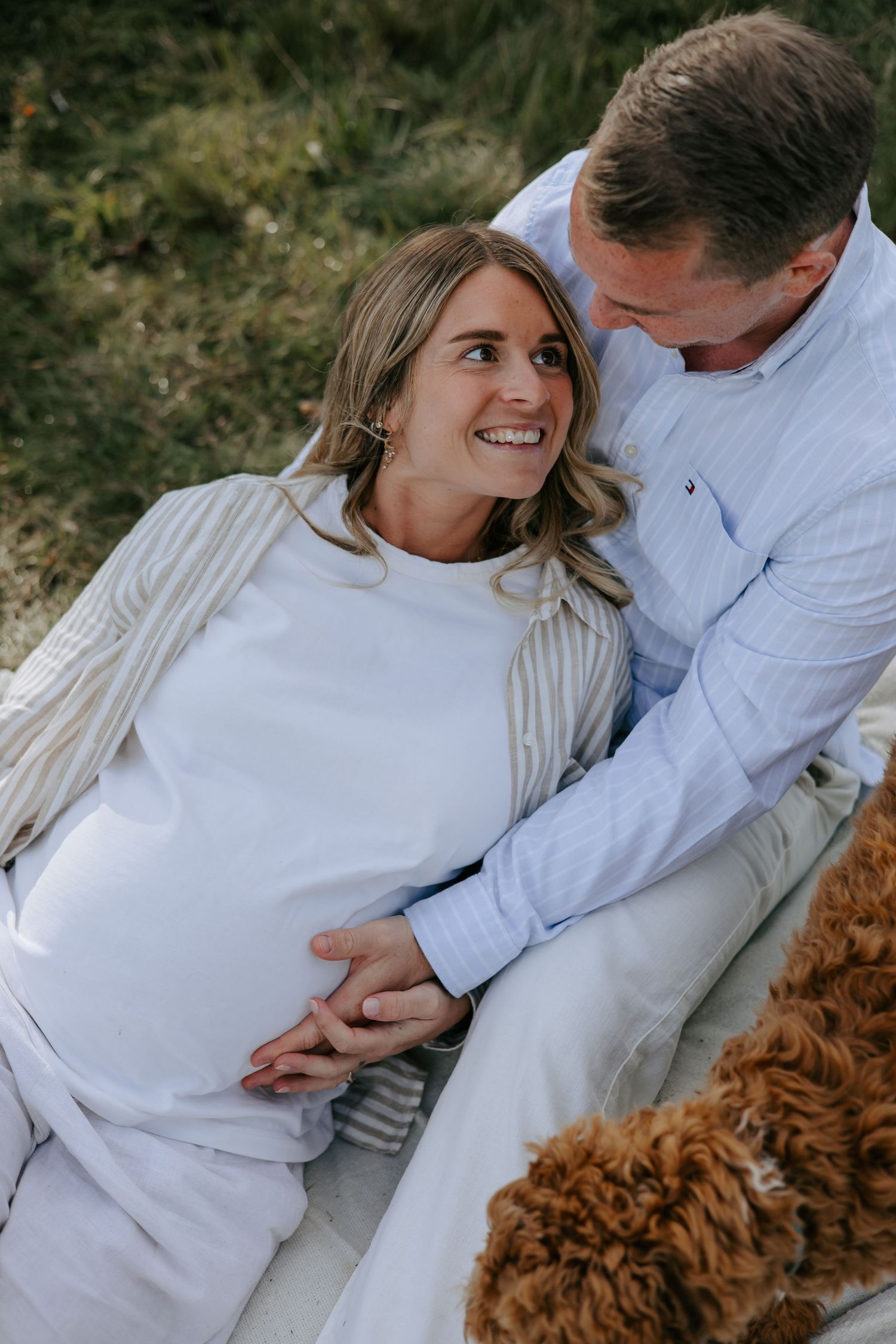 Pregnant woman smiling at partner outdoors on blanket with fluffy brown dog, maternity photo session in nature.