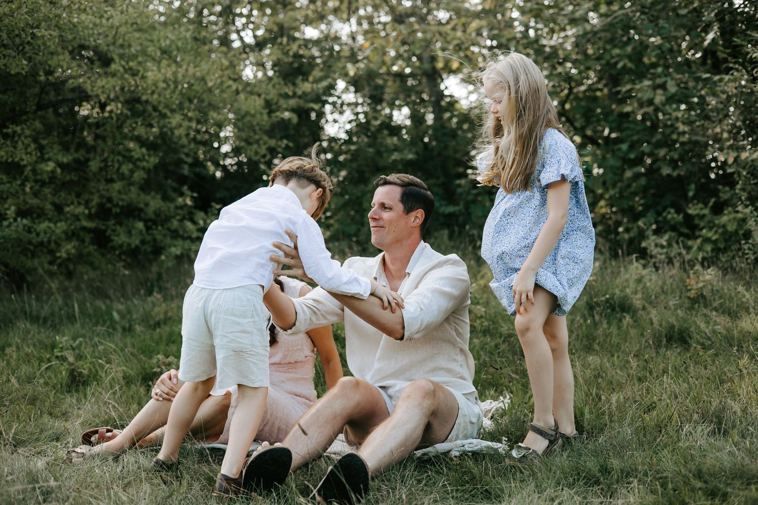 Father sitting on grass playfully interacting with two young energetic children outdoors.