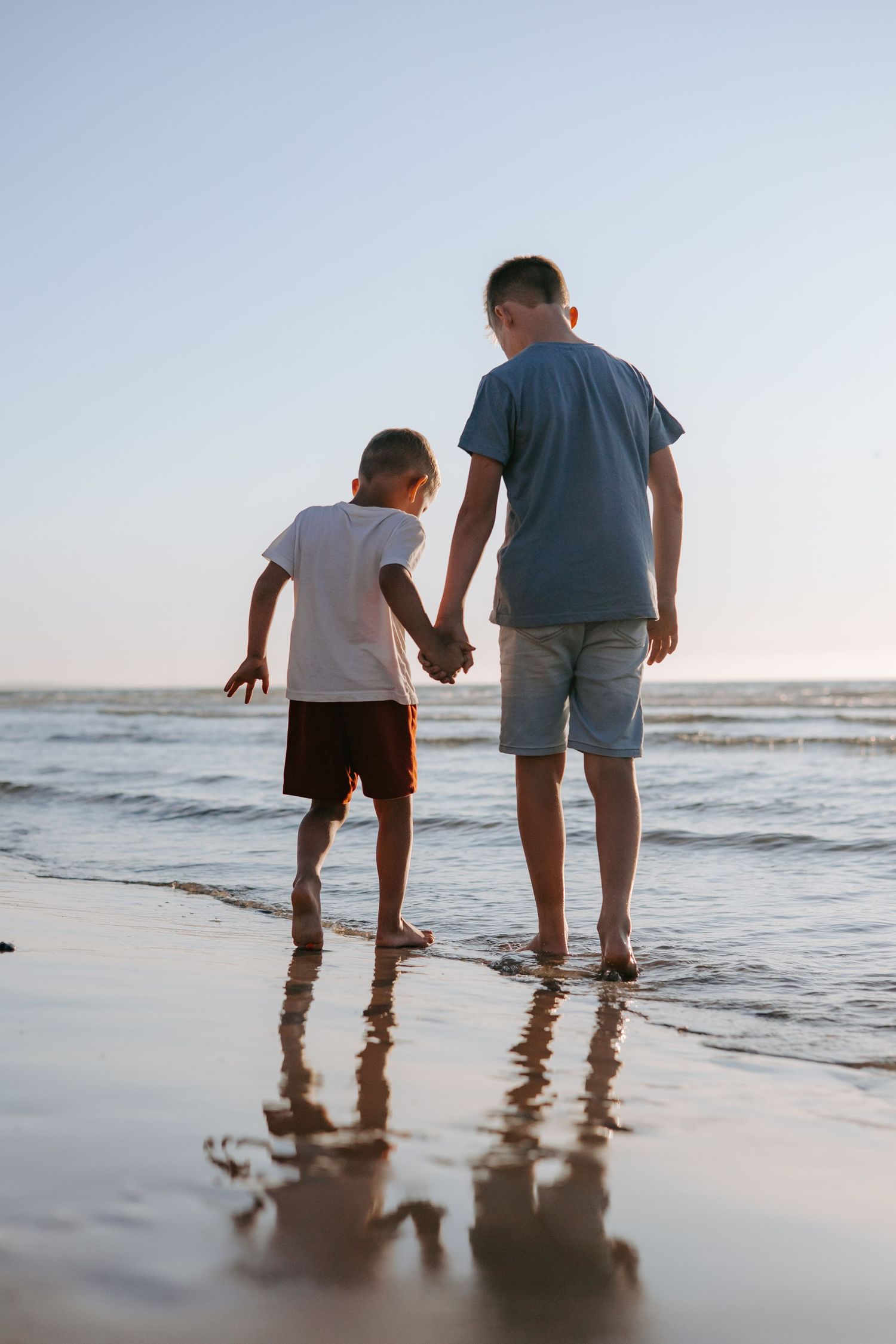 Two boys holding hands while standing barefoot at the ocean's edge during sunset on a sandy beach.