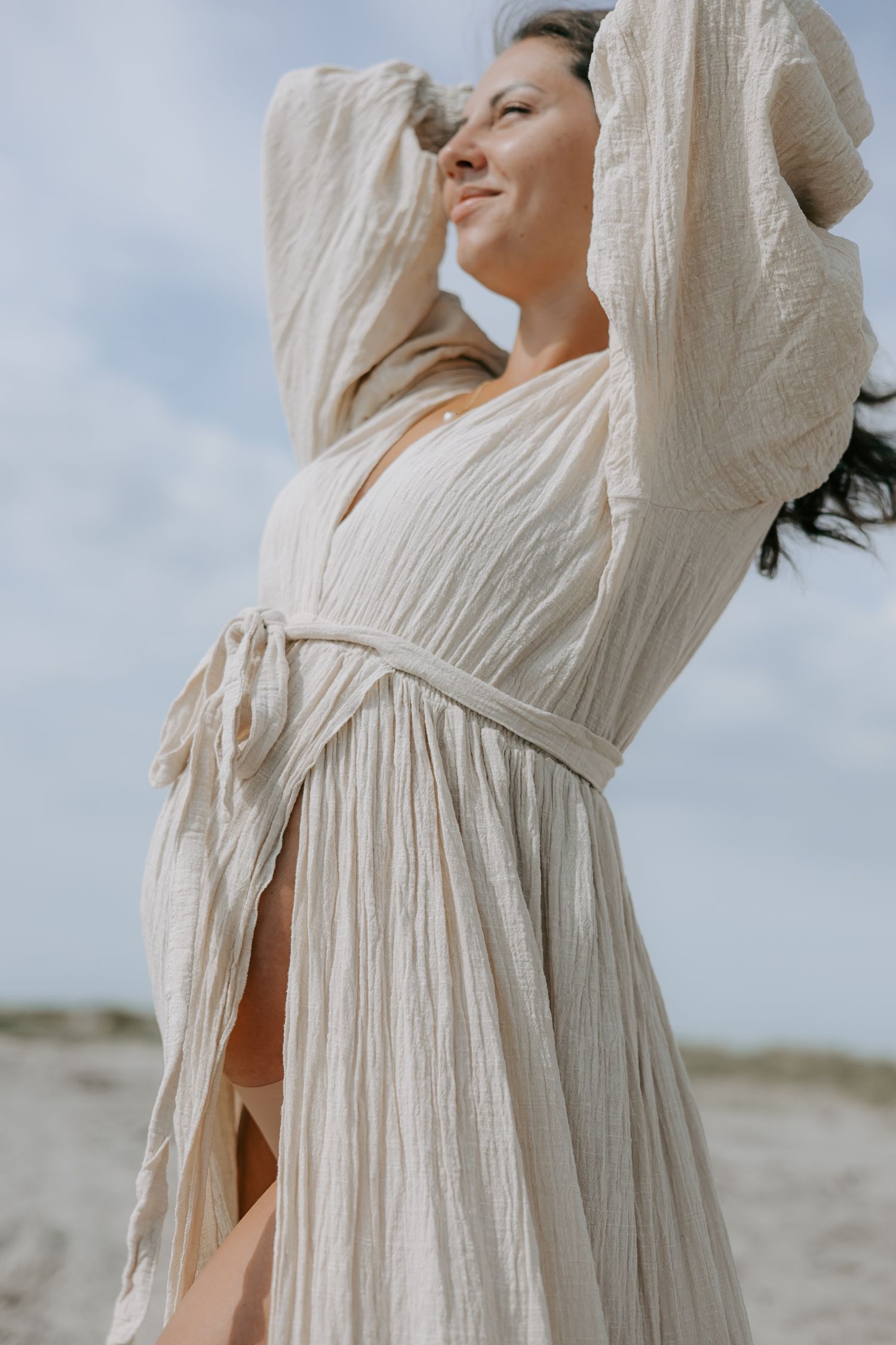 Smiling pregnant woman in beige textured wrap dress posing on sandy beach.