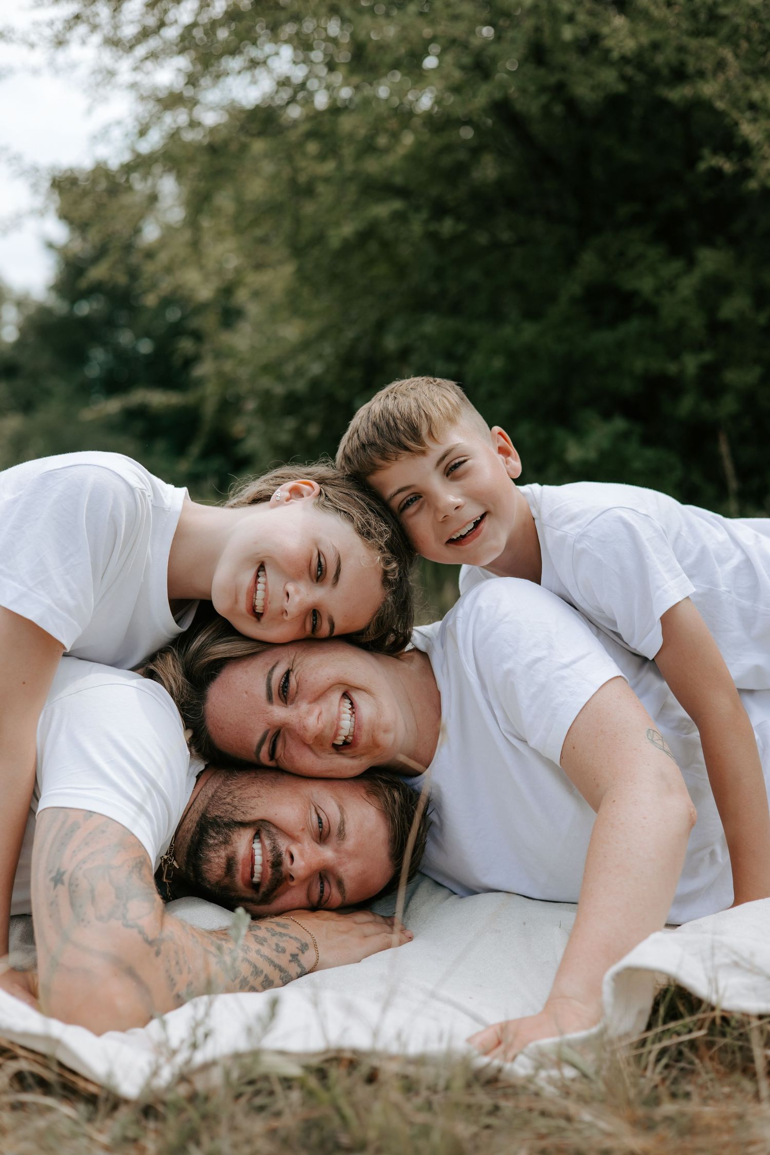 Happy family of four in white t-shirts stacking heads outdoors on blanket in nature.