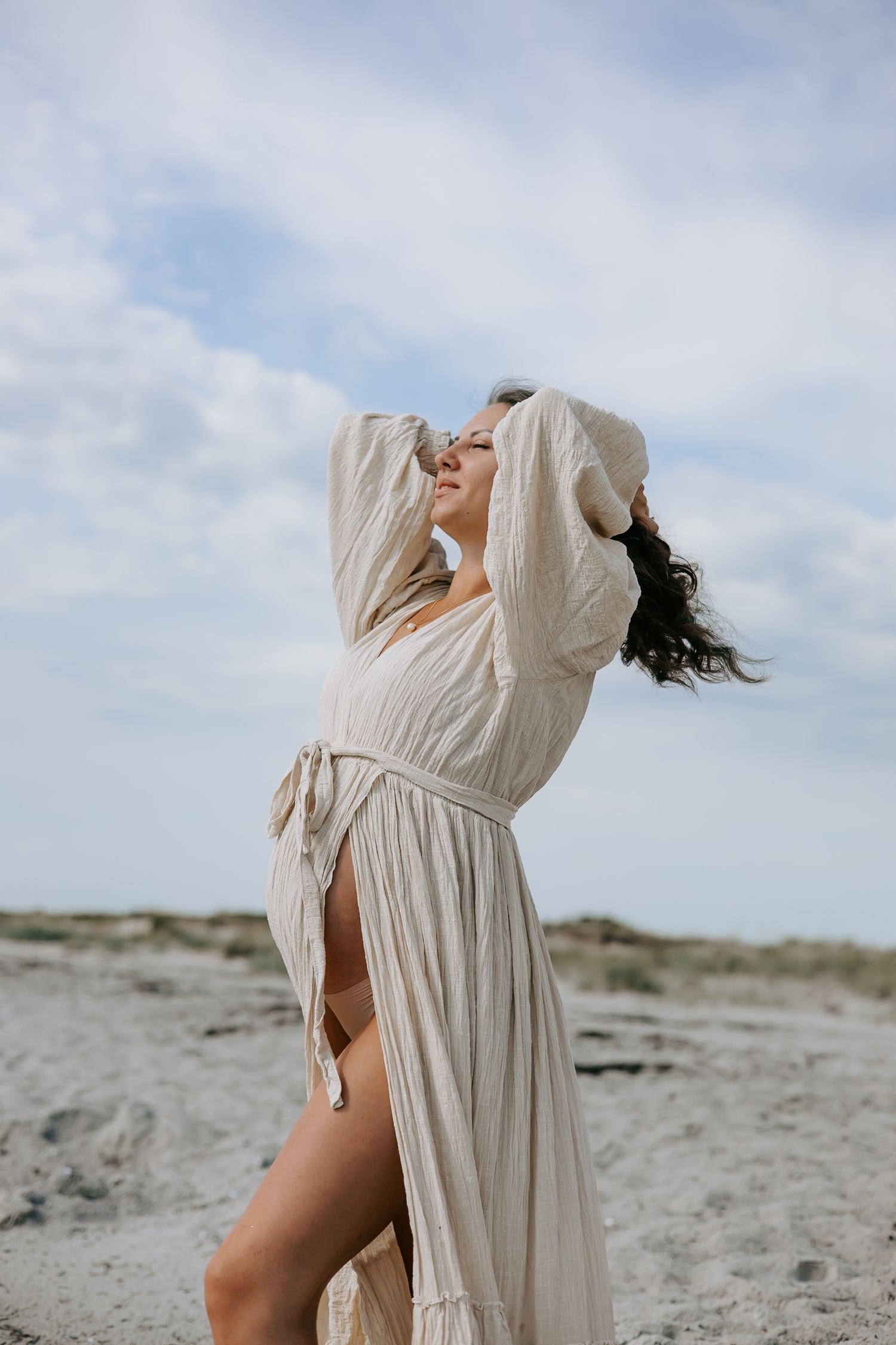 Pregnant woman in flowing cream dress with hands in hair on sandy beach.