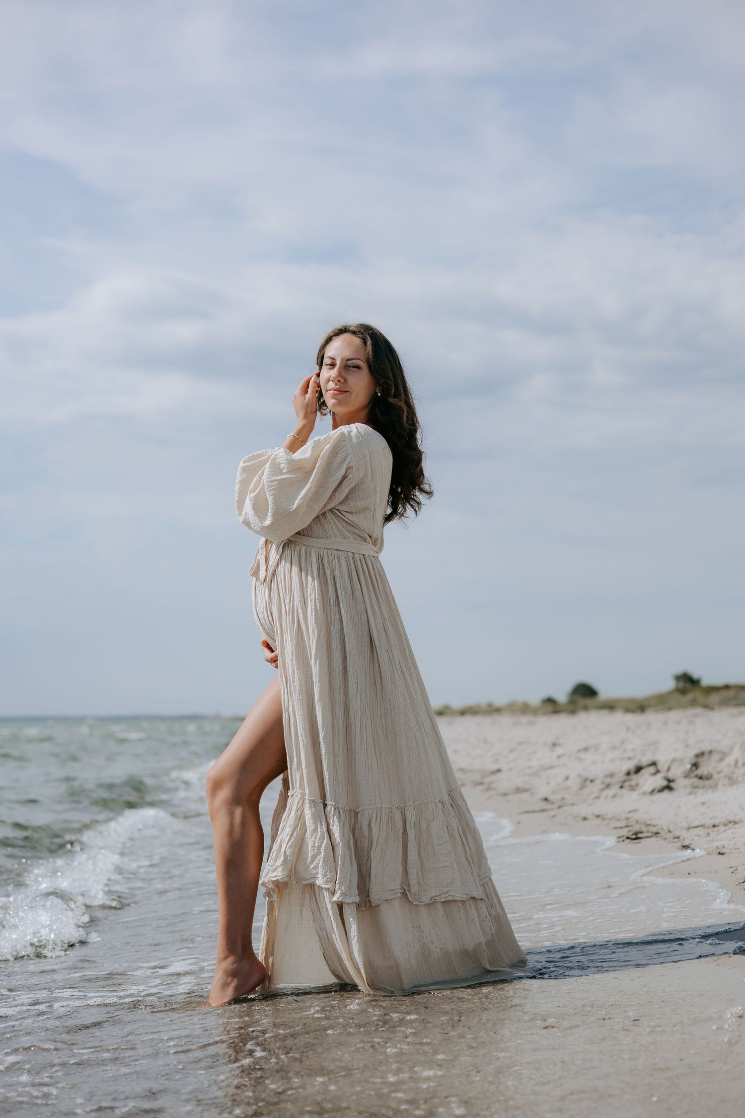 Pregnant woman in flowing beige dress posing gracefully on sandy beach by ocean.