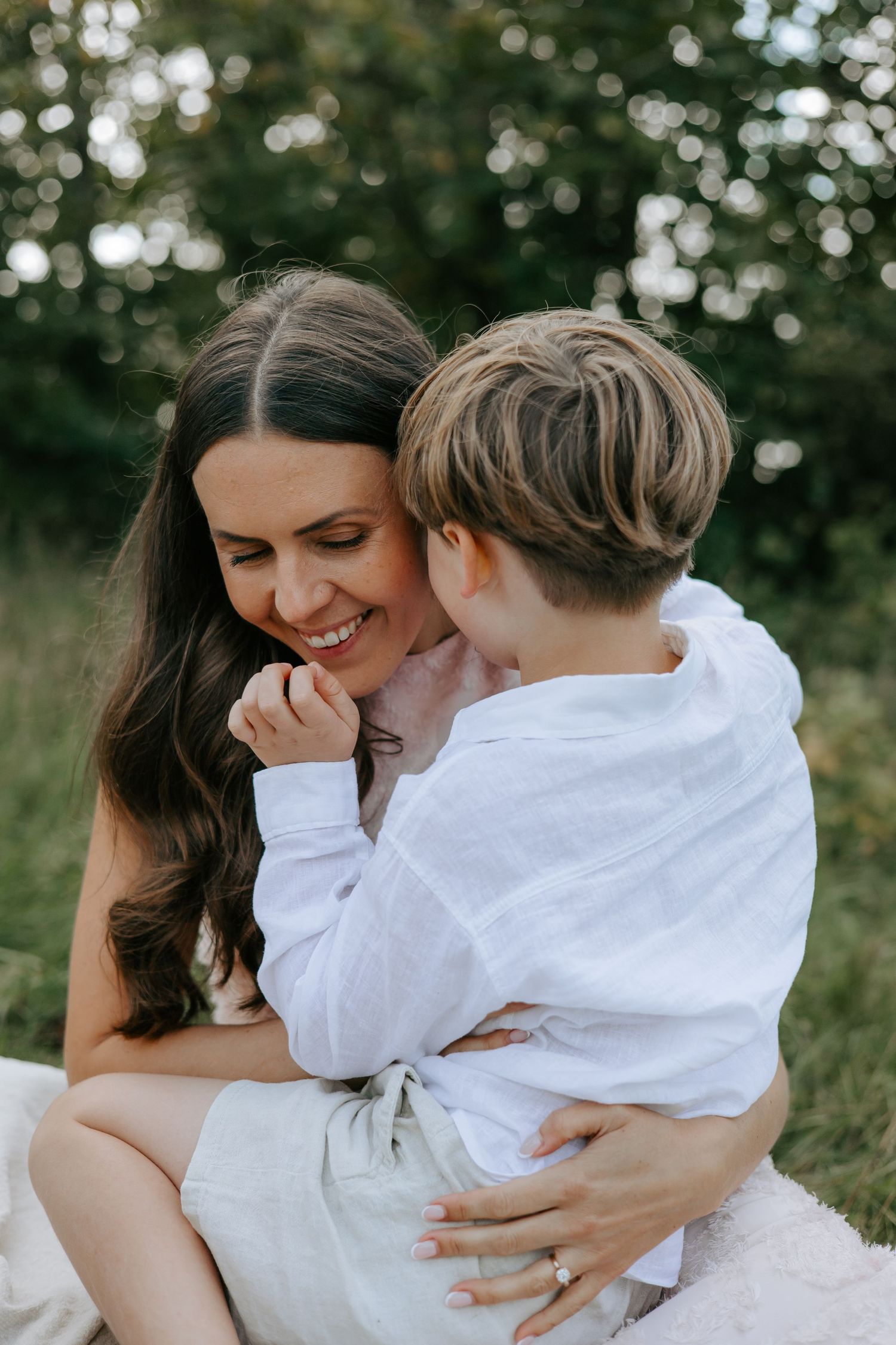 Laughing mother embracing young son on her lap in green field.