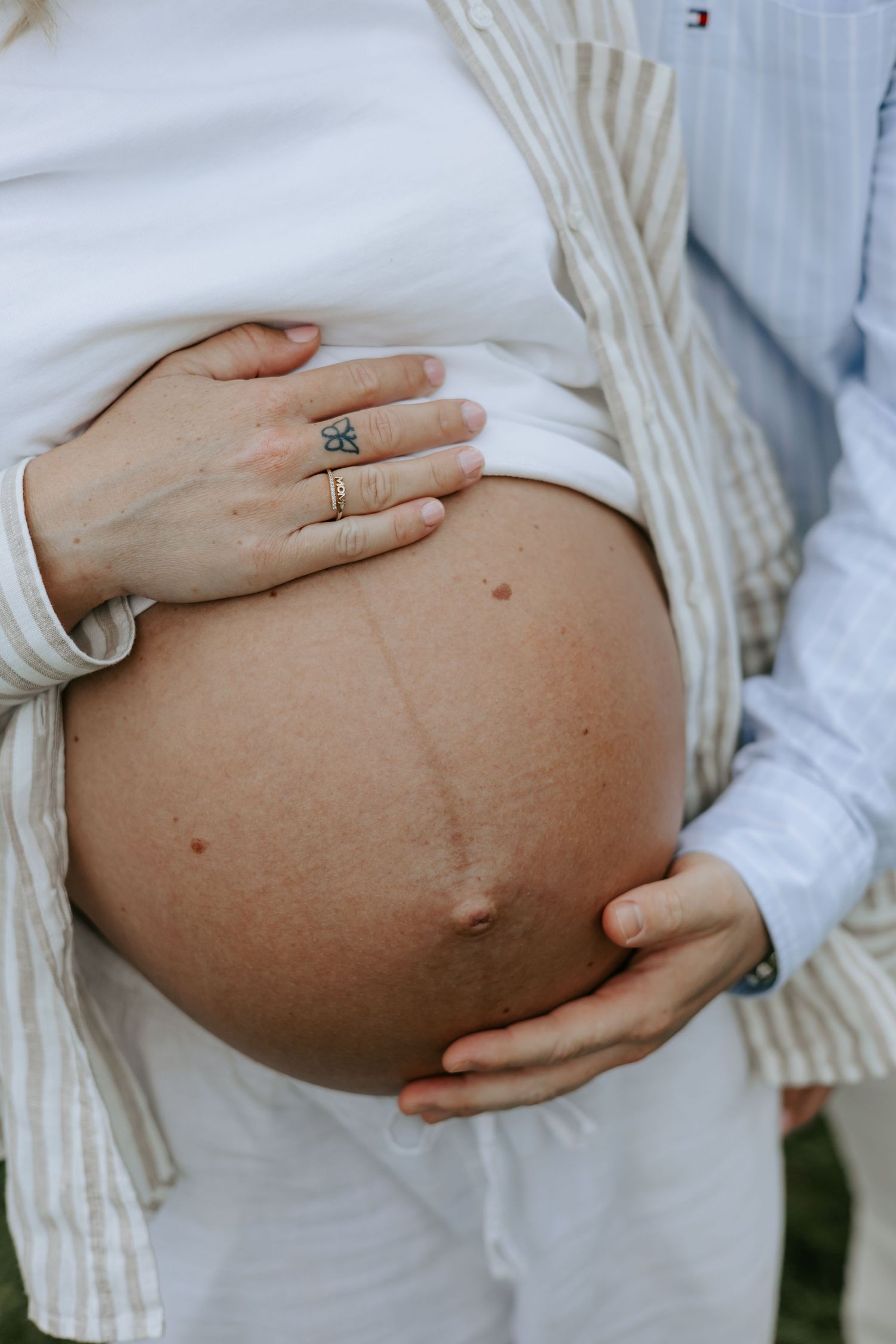 Close-up of pregnant woman's bare baby bump with hands cradling it, wearing rings and white outfit outdoors.