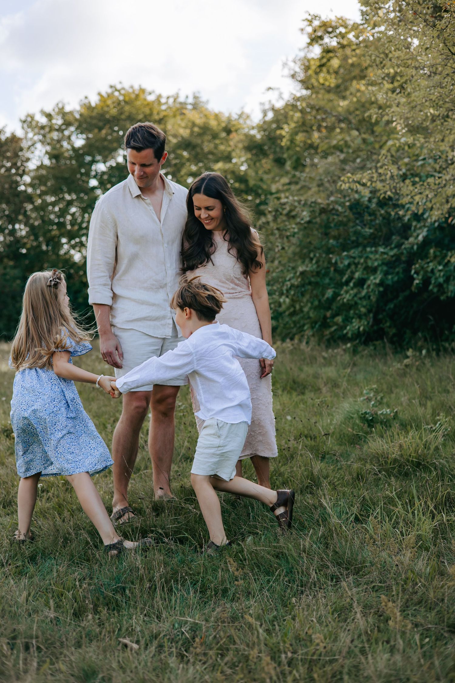 Family of four playing together in a sunny green meadow, parents smiling at children holding hands.