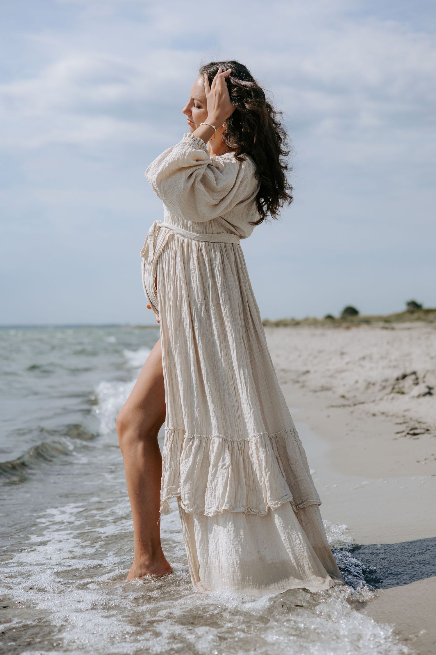 Pregnant woman in flowing beige linen dress standing in ocean waves on sandy beach.