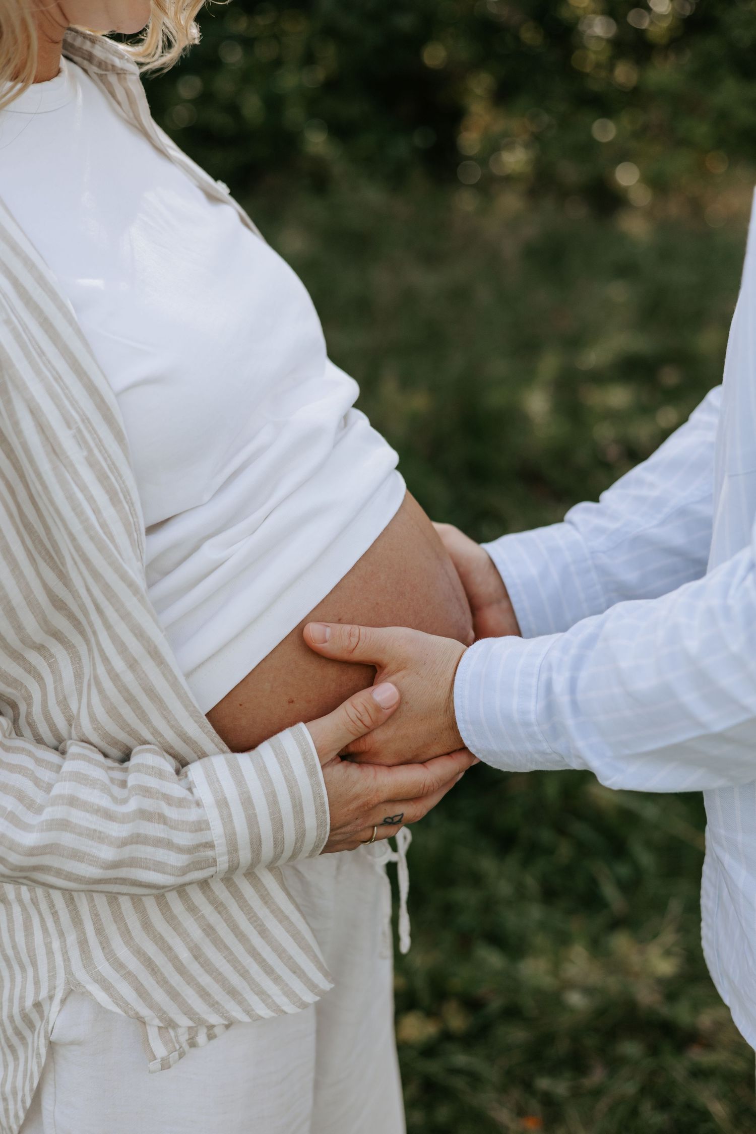 Couple holding pregnant belly outdoors, wearing white and beige linen outfits, surrounded by lush green foliage.