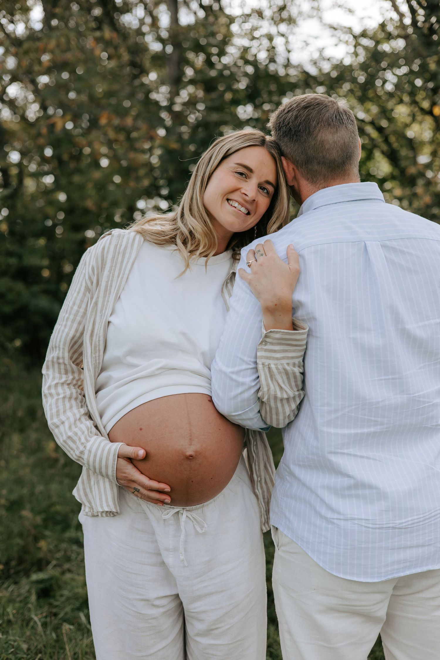 Smiling pregnant woman in white outfit holds her baby bump while partner kisses her forehead outdoors.