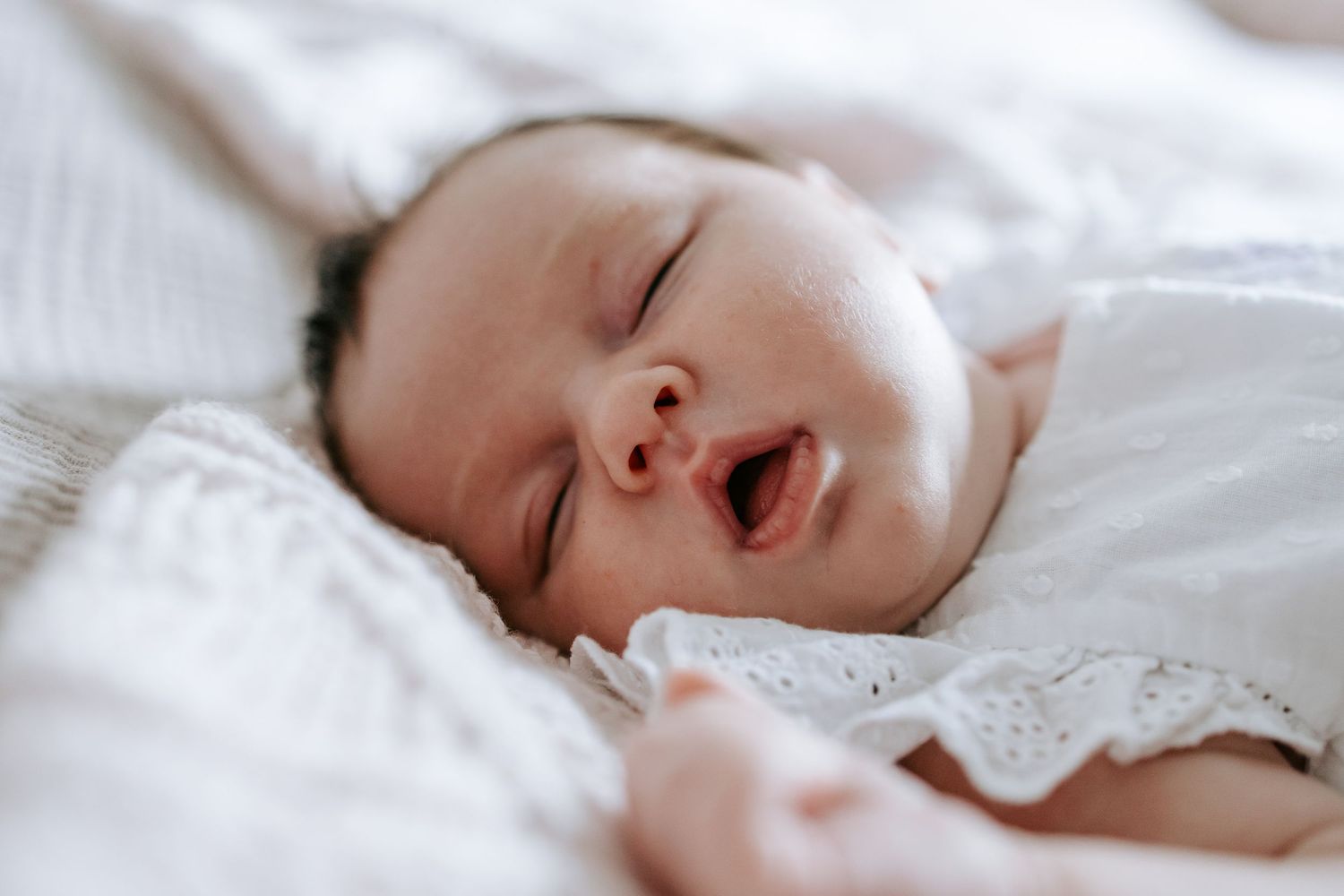Peaceful newborn baby sleeping with mouth open on white soft blankets.