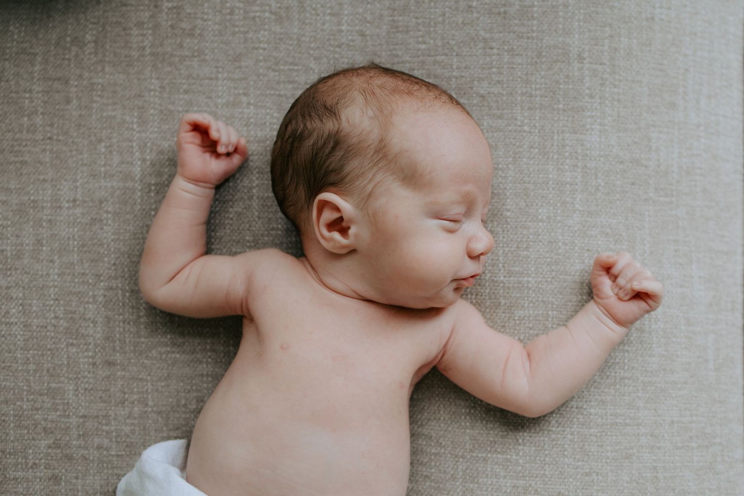 Peaceful newborn baby sleeping on a beige surface with arms raised up.