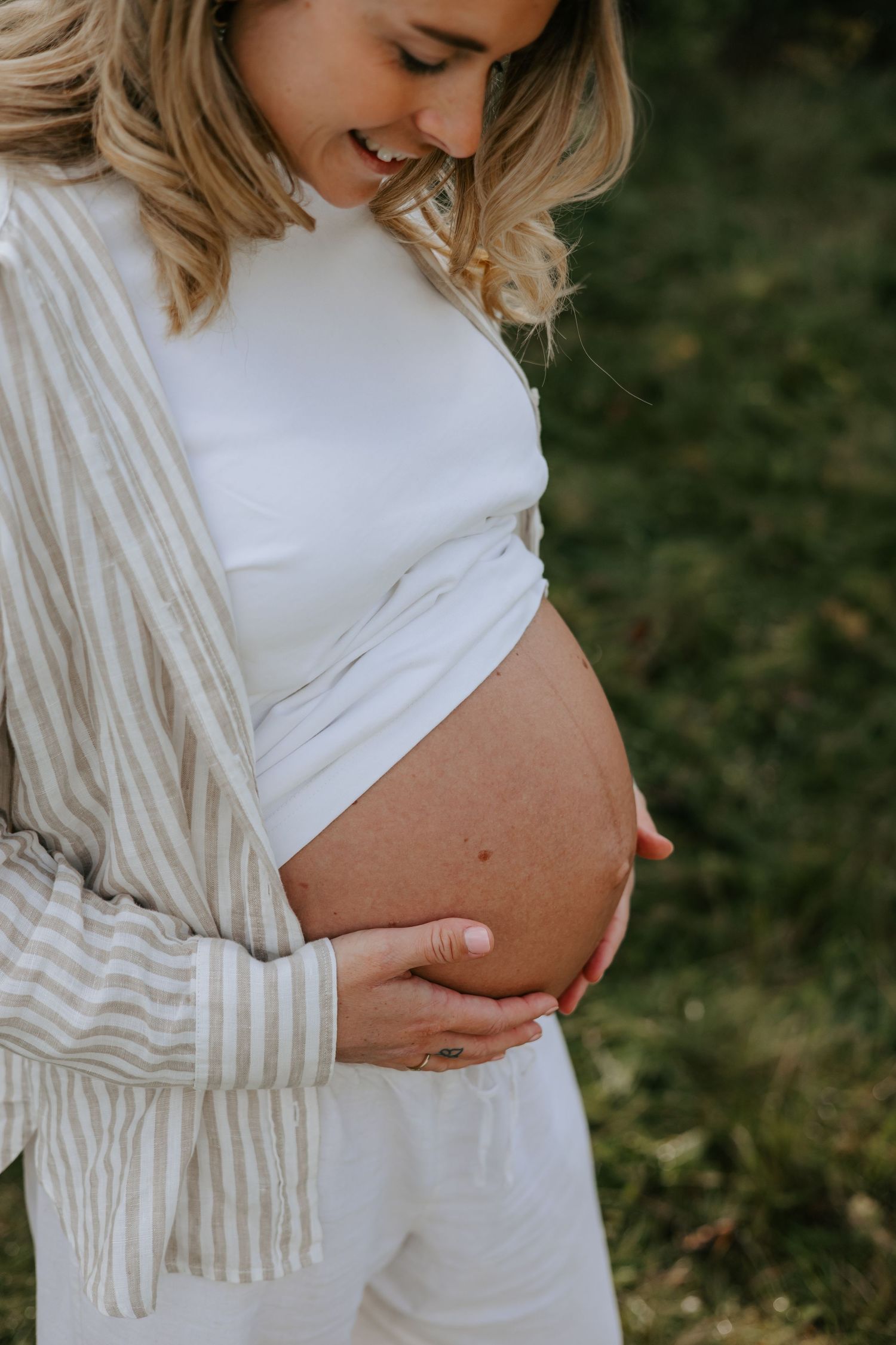 Smiling pregnant woman in white crop top and striped shirt cradling her baby bump outdoors in natural light.