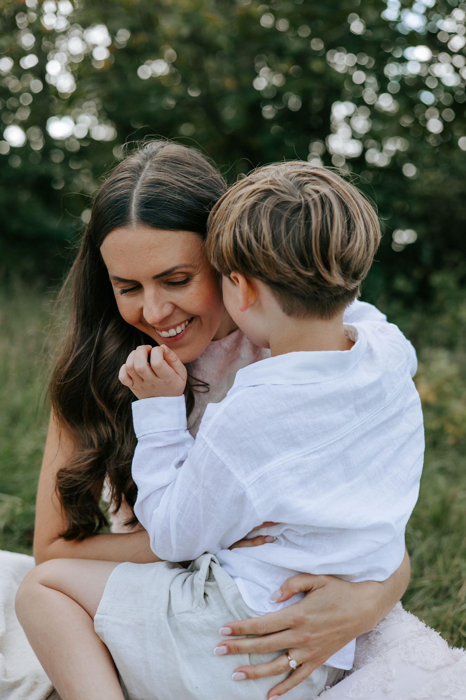 Smiling mother hugging her young son outdoors in a green park setting.