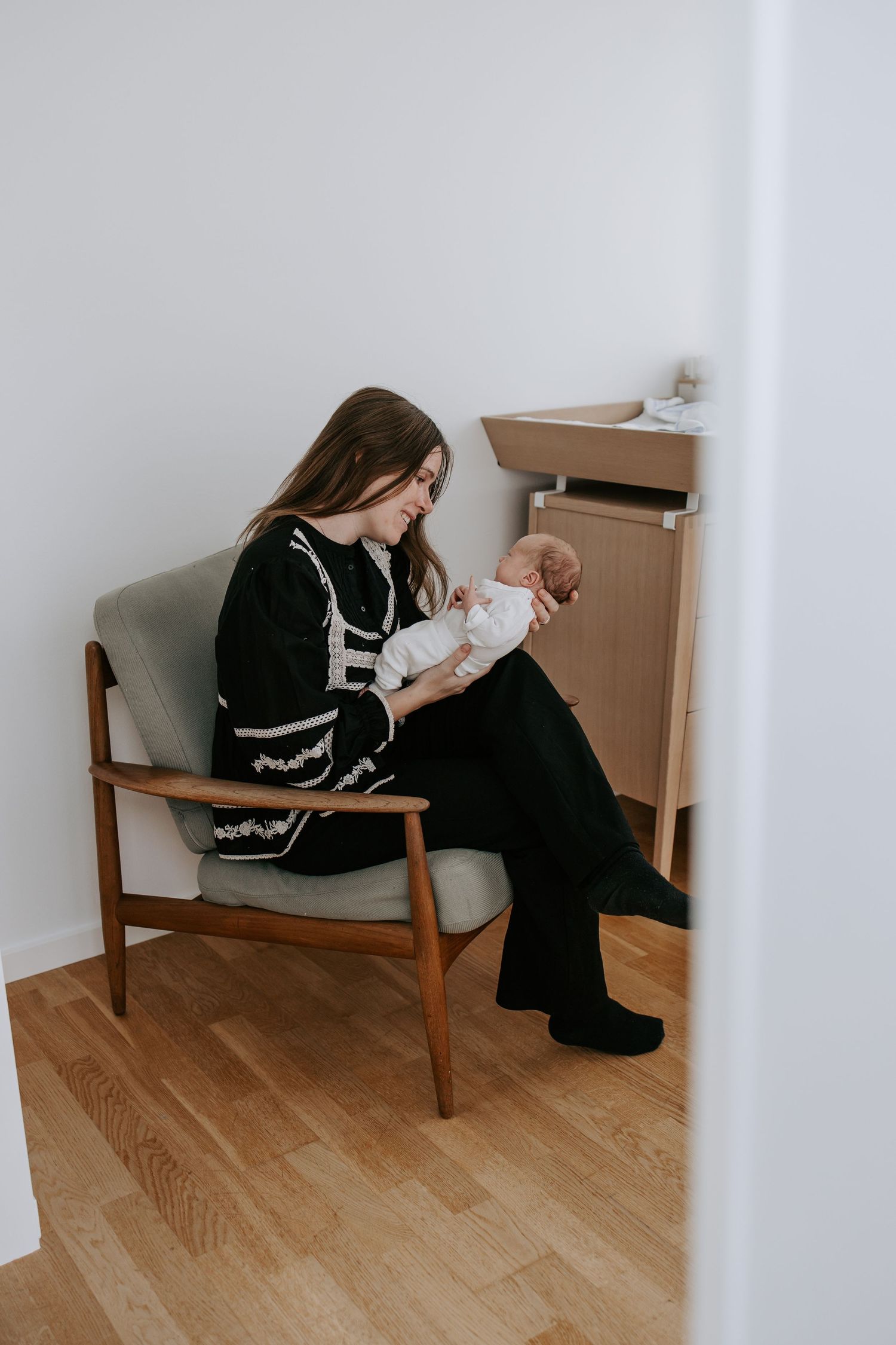 Mother in black dress cradling newborn baby in wooden armchair at home.