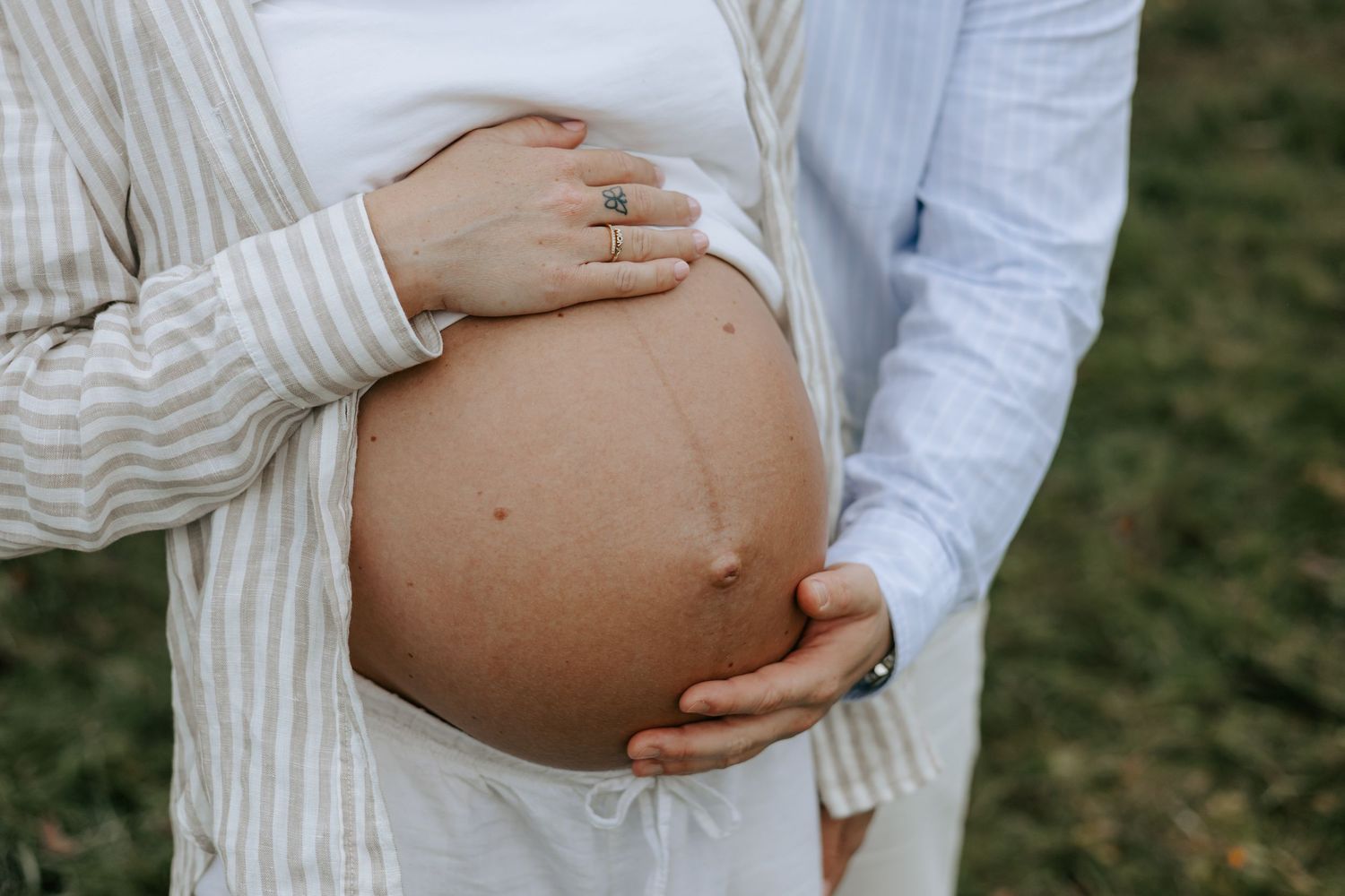 Pregnant woman in striped cardigan with partner's hand on bare baby bump outdoors.