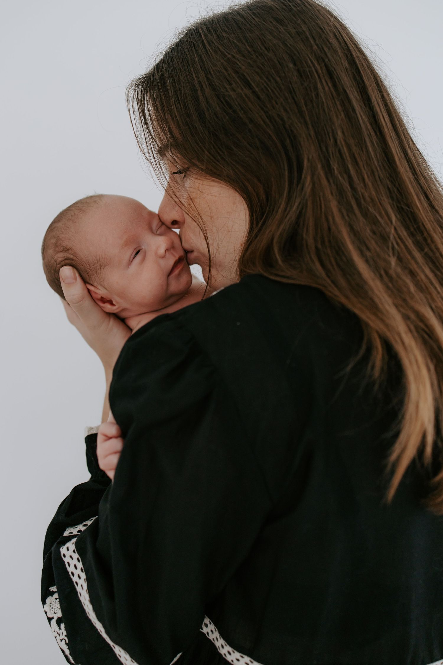 Mother with long brown hair tenderly kissing her sleeping newborn baby.