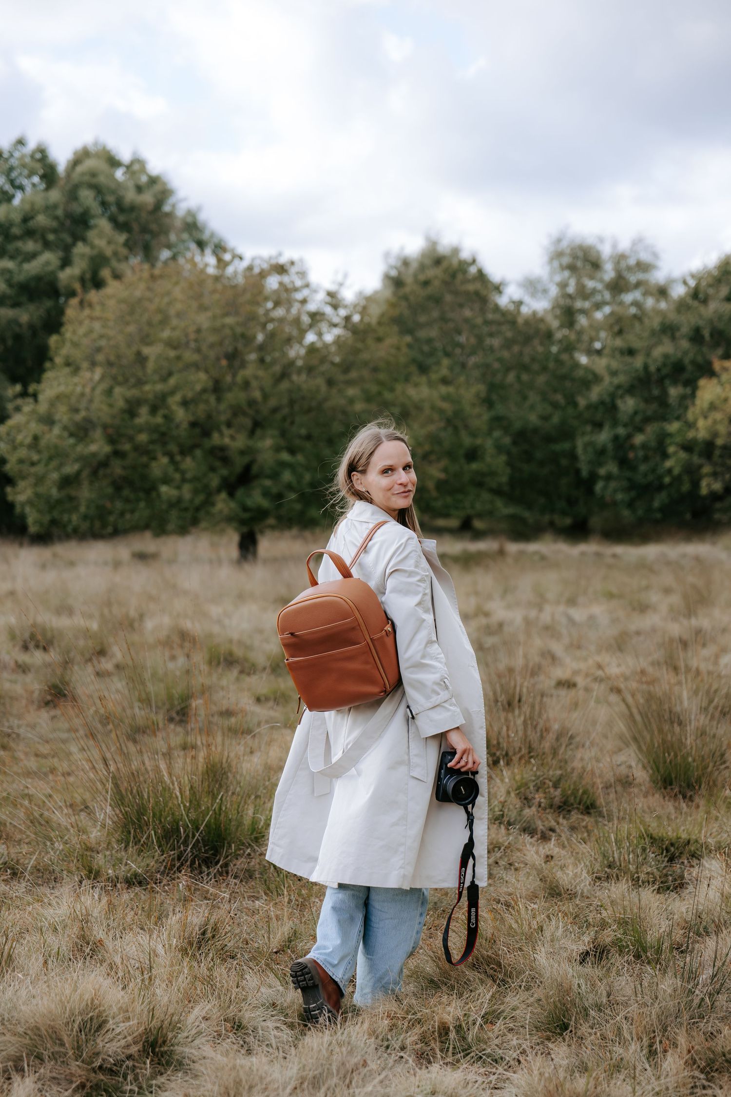 Woman in white coat with brown backpack holding camera stands in dry grass field with trees in background.